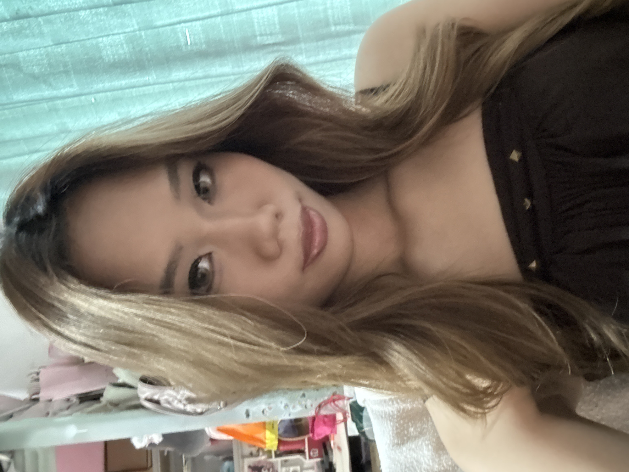 A woman with long, light brown hair is lying down and looking at the camera. She is wearing a dark brown top. The background includes a blue curtain and what appears to be a cluttered area with clothes and shelves. The image does not contain sufficient information to pinpoint the location in the city of Nan'an District, China.