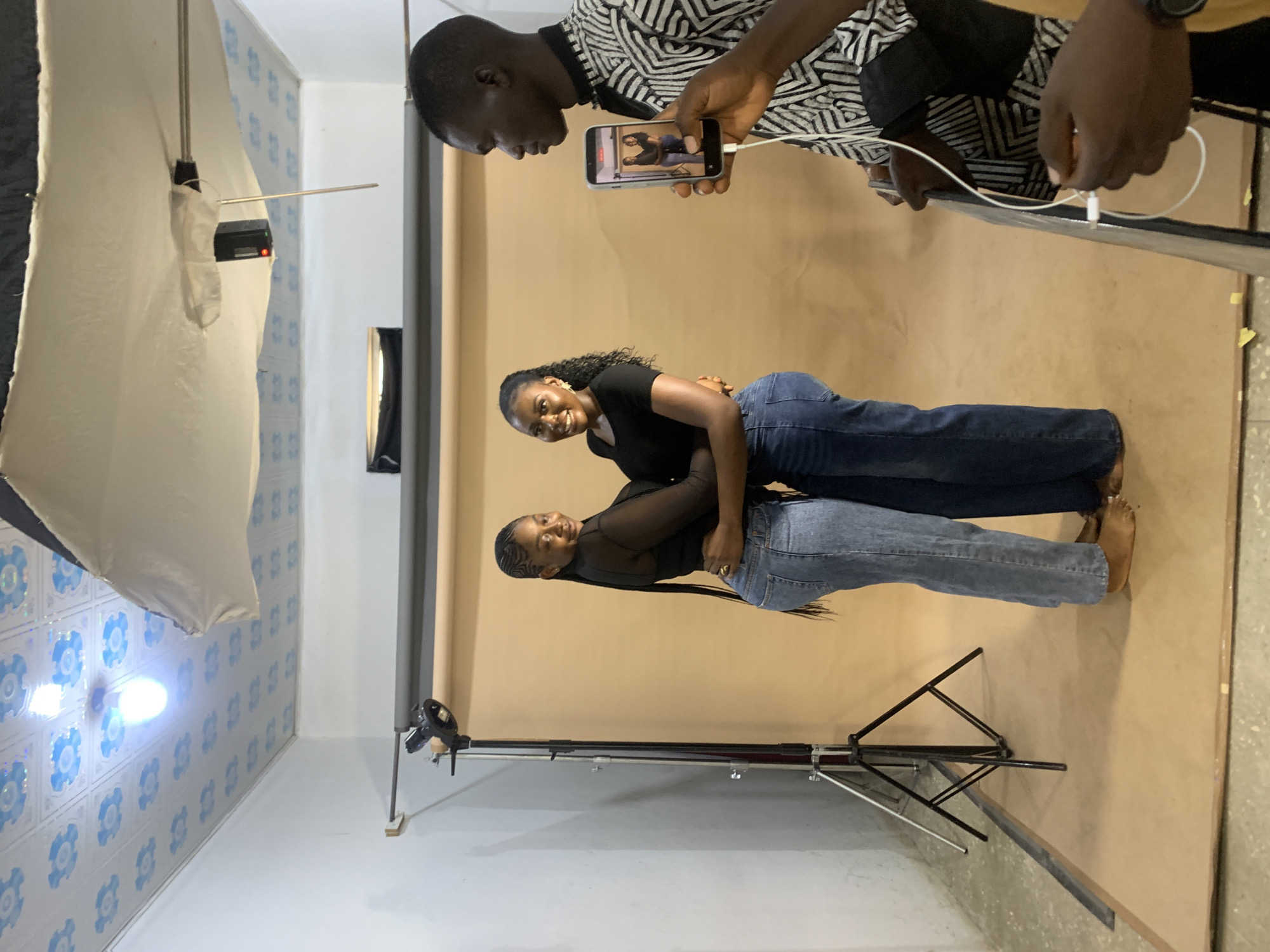 In Gura Top, Nigeria, a man films two young women posing for a photograph. The women, dressed in black tops and denim jeans, stand close together in front of a tan backdrop. A professional lighting setup, including a large diffused light and a smaller spotlight, illuminates the scene. The man, wearing a black and white patterned shirt, holds up a smartphone to capture the moment, displaying a preview of the photo on its screen. The room has a white-tiled ceiling with floral designs and a single lightbulb. A camera tripod is visible to the left of the subjects. The floor is a smooth, textured surface.