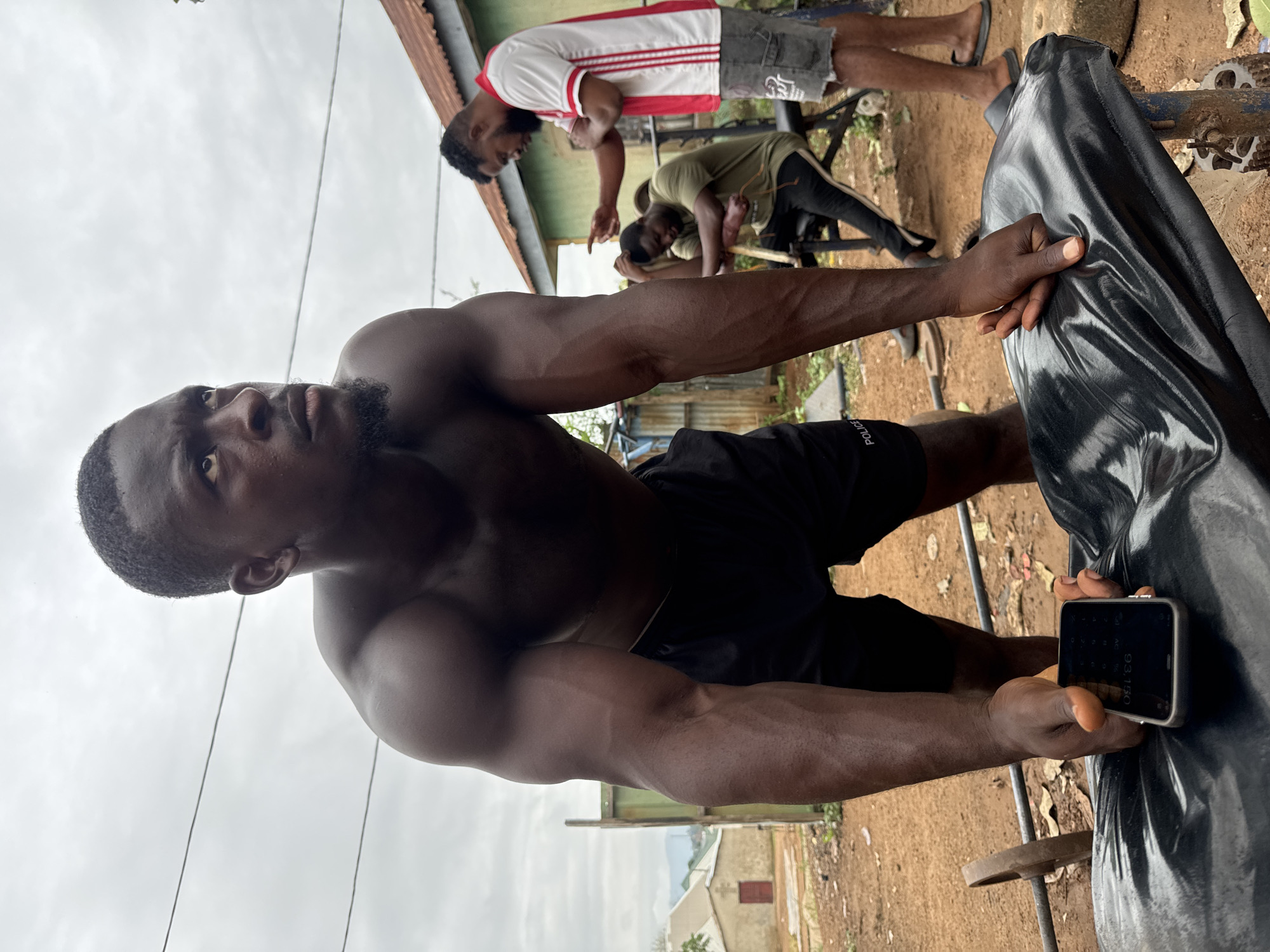 In Akwanga, Nigeria, an outdoor gym scene unfolds under an overcast sky. A shirtless Black man, with dark skin and a muscular build, is the central focus. He is positioned in the foreground, leaning over a black padded bench, his hands resting on it as he looks upwards and to the right with a determined expression. A smartphone, displaying a calculator app with the number "93.150", is held in his left hand. He wears black shorts with "POLICE" written in white on the leg.
Behind him, two other Black men are visible. One, wearing a green t-shirt and black pants, is seated on a weight bench, looking down. The other, standing and gesturing with his right hand, is wearing a white t-shirt with red stripes and grey shorts. The environment is rustic, with reddish-brown earth, scattered dry leaves, and some vegetation. A corrugated metal fence and simple buildings with reddish roofs are in the background, suggesting a residential or semi-rural area within Akwanga. A weight bar with a disc is partially visible on the left. The overall atmosphere conveys a sense of focused physical training in a modest, communal setting.