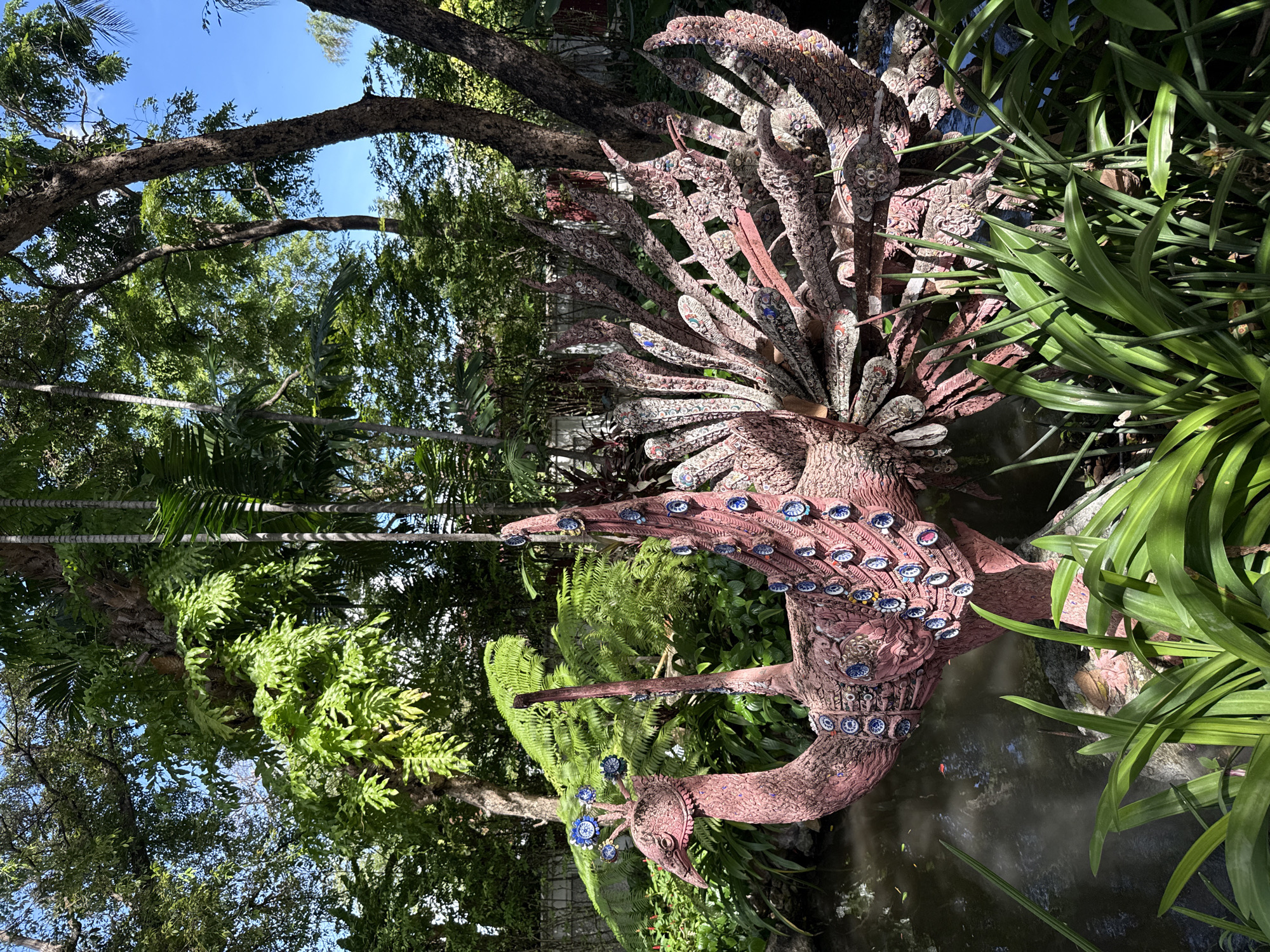 A mosaic-covered peacock sculpture stands within a garden setting in Samrong Nuea Subdistrict Municipality, Thailand. The sculpture is pinkish-red, adorned with blue and white circular mosaics that resemble eyes along its neck and tail feathers. The peacock is positioned near water, potentially a pond or small stream, surrounded by lush green vegetation, including various plants and trees. The scene suggests a garden or park setting, likely a tourist spot. The photo is shot with a wide-angle lens under natural sunlight, casting shadows and highlighting the textures of the mosaic and the foliage.