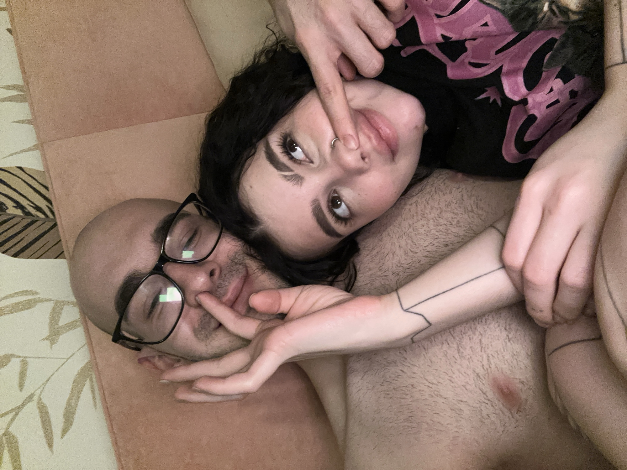 A photograph depicts two individuals lying together on a peach-colored, cushion-like surface. One person is bald and wearing glasses, lying on their back. The other person, with dark curly hair and a nose ring, is lying on top. One hand is manipulating the face of the person on top, while the other hand is entangled with the person on the bottom. The setting is presumably indoors. There is no information in the image that would allow for the identification of the location in the city of Kazan, Russia.