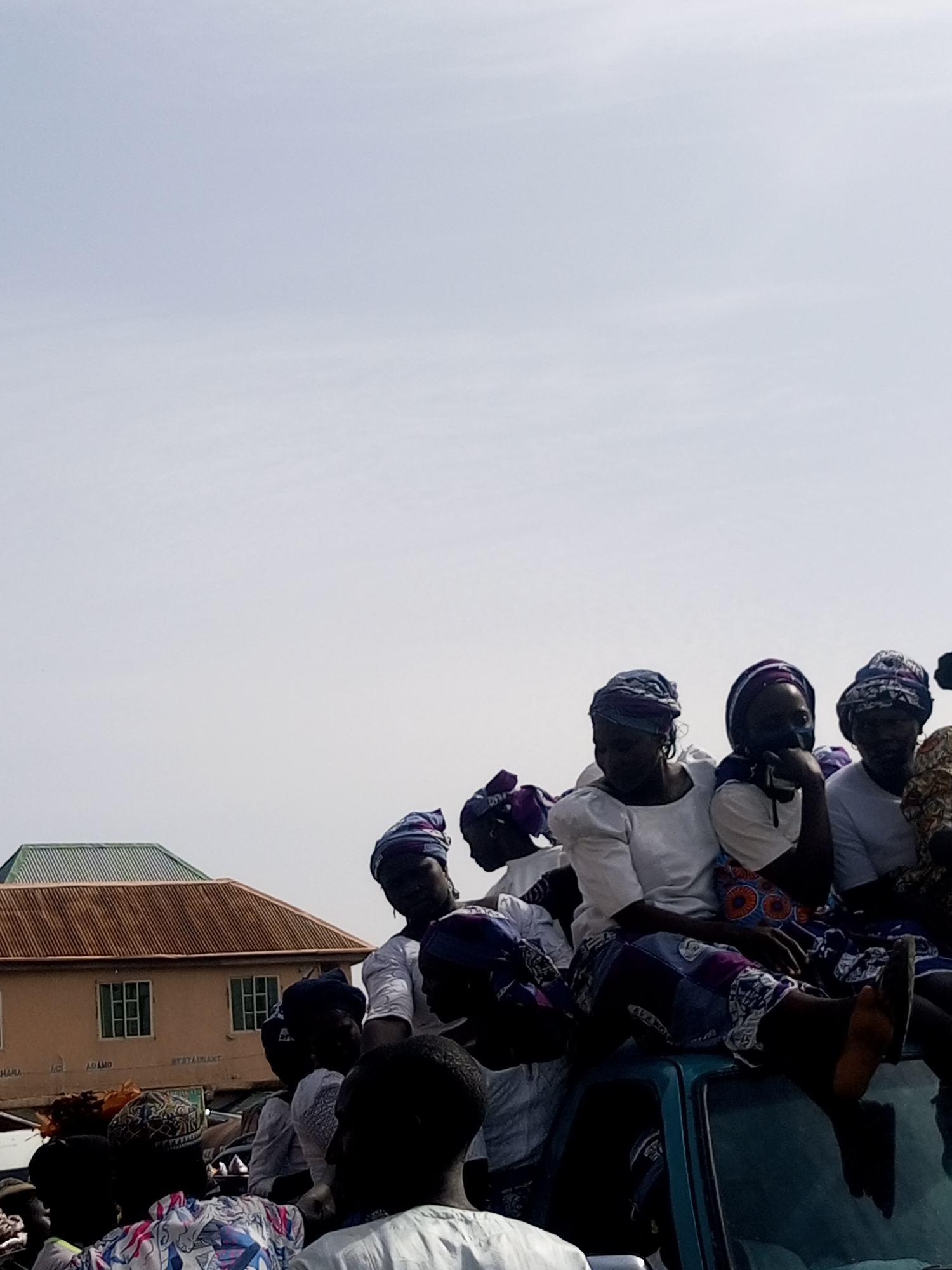 A group of women wearing matching head wraps and primarily white tops are gathered on the roof of a vehicle. The scene appears to be in Gombe, Nigeria, based on local businesses visible in the background, specifically on a building with a brown roof. The women are seated and appear to be interacting with others out of frame, potentially as part of a procession or public gathering. The day is clear and bright.