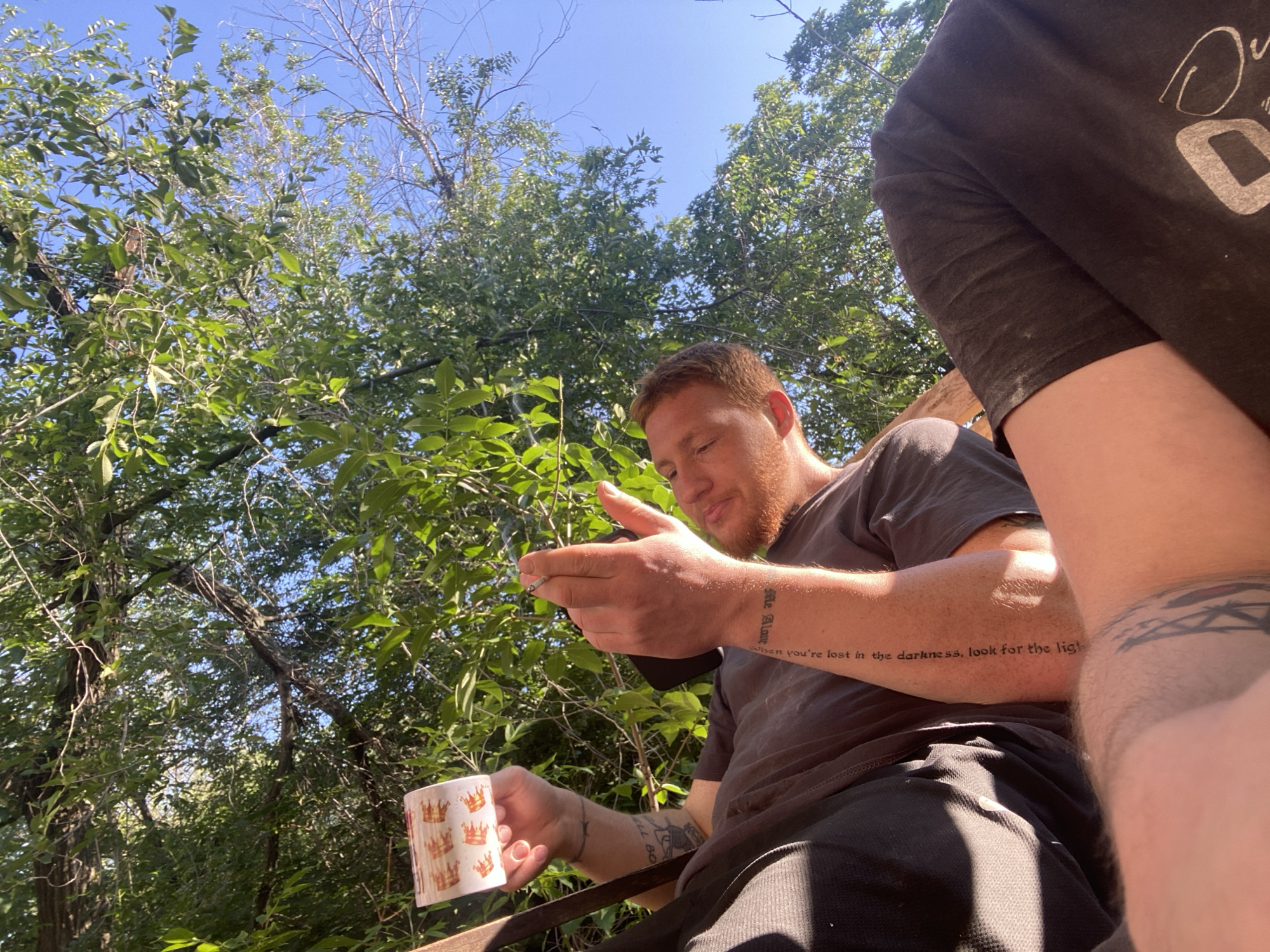 In an outdoor environment with abundant foliage and a clear blue sky, two individuals are partially visible. The foreground features a person holding a white mug with a crown pattern. This individual has ginger hair and is looking down at their hands, which are holding a cigarette. A tattoo is visible on their left forearm. To the right, another person's arm and shoulder are visible, wearing a dark t-shirt with a white graphic. Both individuals appear to be seated.