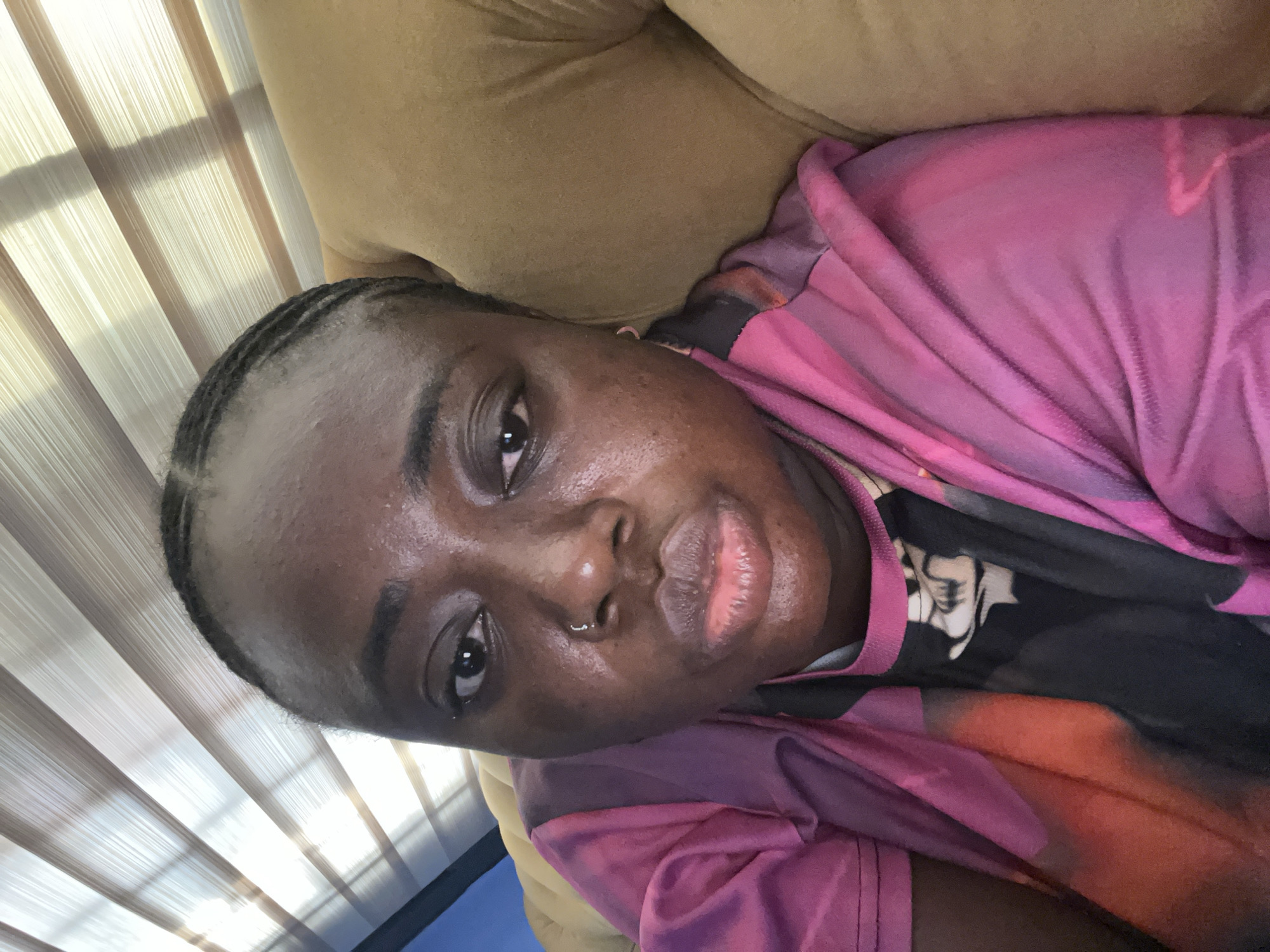 A close-up shot of a woman with dark skin, wearing a pink and black patterned shirt, reclining on a tan sofa. She has short, dark hair styled in braids, and a silver nose ring on her left nostril. The background includes a window with vertical blinds. There is no evidence to suggest this image was taken in Kubwa, Nigeria. Location is unknown.