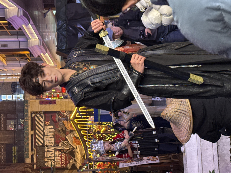 An East Asian male in a black, multi-layered costume stands centrally in an urban, illuminated setting in Yuzhong District, China, at night. He wears a dark headband with a red ornament and features three small golden-orange markings on his right cheekbone. An ornate necklace with red, blue, and gold elements is visible on his chest. He holds a silver-bladed sword with an ornate hilt in his right hand, partially drawing it from a black, gold-accented scabbard held in his left hand. He gazes forward with a slight smile.
To his left, a young East Asian female in traditional attire stands on a raised platform, wearing an elaborate silver headdress, a dark top with red embroidery, and a dark skirt, holding a dark, ornate umbrella-like object. Behind her, two or more women in ornate dresses are partially visible. In the mid-ground to the right of the central male, an older East Asian male in a blue cap and dark jacket holds a camera, facing the subjects. A person wearing a wide-brimmed straw hat and dark jacket is seen from behind, crouching in the lower mid-ground. The lower right foreground shows the shoulder and head of another individual in a light-colored puffer jacket.
The background features illuminated building facades. To the left, a large wall mural depicts a mecha-style robot figure (reminiscent of Evangelion's EVA unit) with Chinese characters and "SMC" visible, adorned with yellow string lights. Arches of yellow string lights are visible above the female in traditional dress. Above and to the right of the central male, a building facade with illuminated window frames (purple and orange light) extends upwards. The scene suggests a themed street or entertainment area.