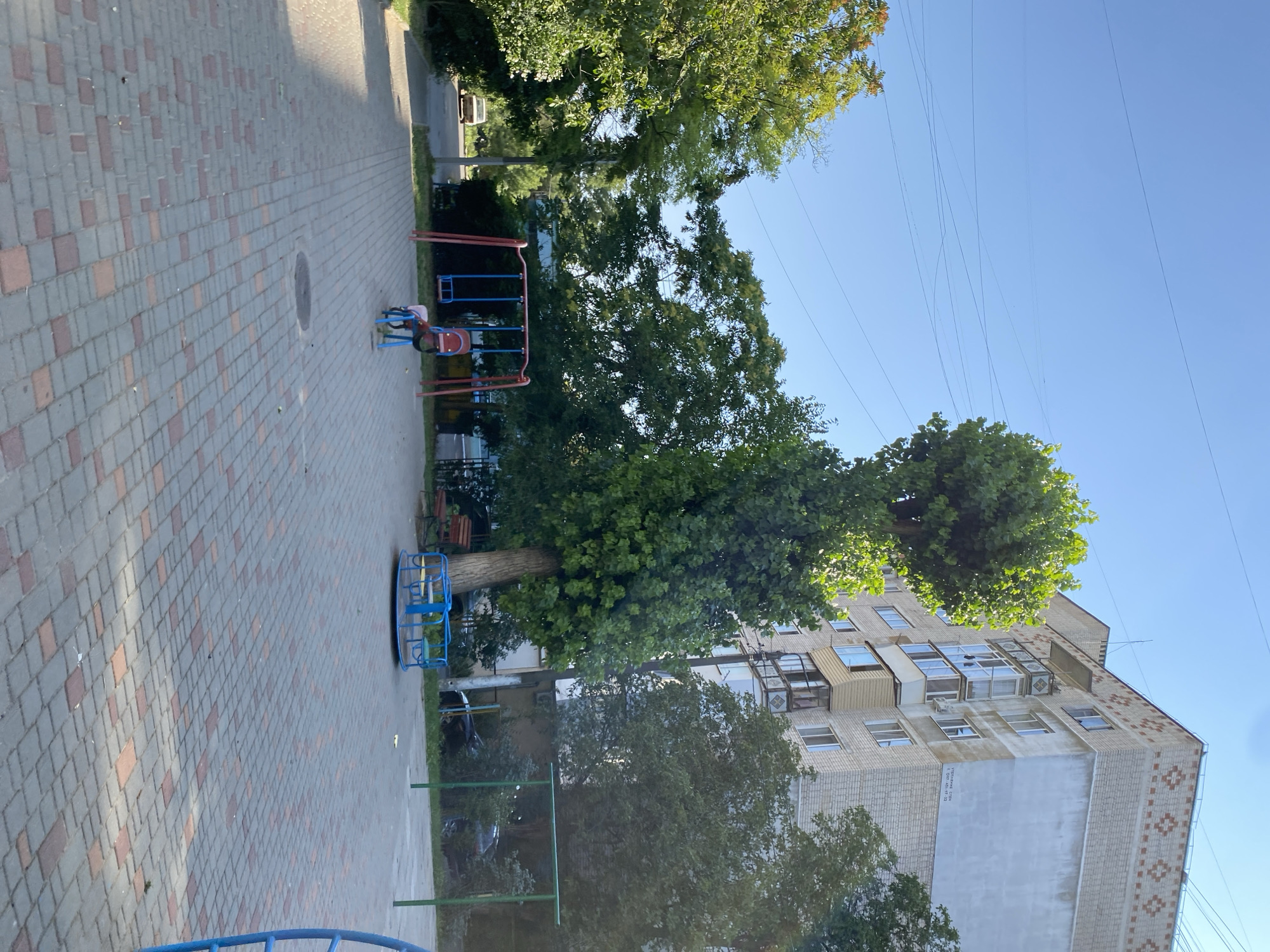 The image shows a children's playground in the city of Chornomorsk, Ukraine. The playground features a swing set with two swings, a merry-go-round, and a child on a toy. In the background, there is a tall, leafy tree, a multi-story apartment building, and some smaller trees. The ground is paved with interlocking bricks. The sky is clear and blue. The scene appears to be set during the daytime, likely in the summer or early fall, given the lush greenery.