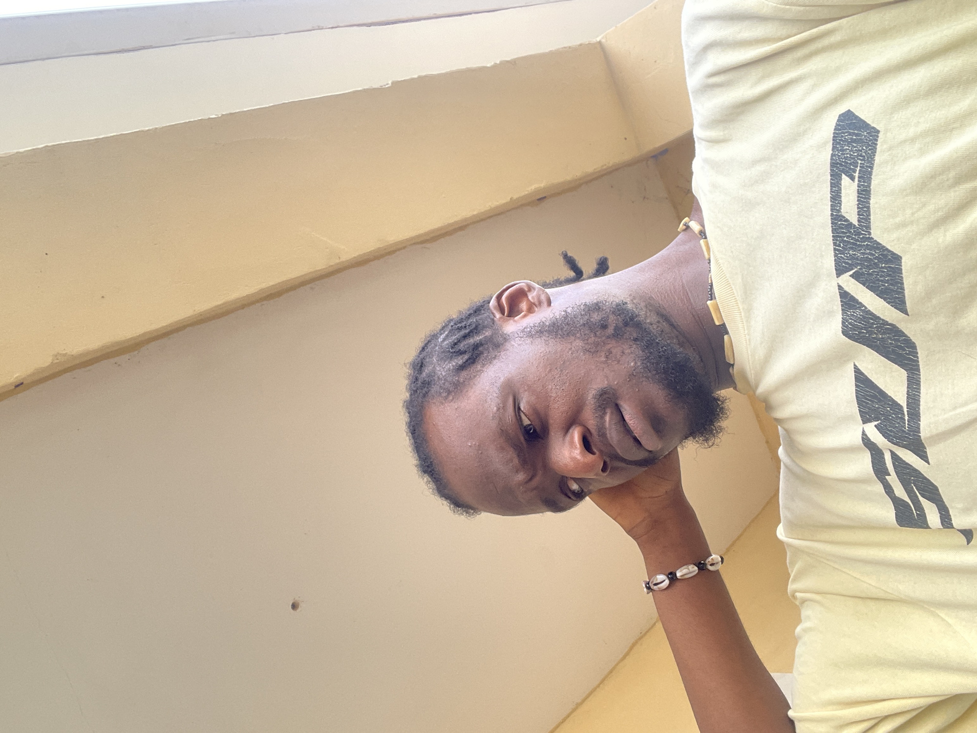 The media file is a close-up, low-angle, portrait-style photograph of a Black man. He is wearing a light yellow t-shirt with a large, dark grey graphic design on the chest. The design appears to be abstract or stylized lettering, possibly an emblem or logo. He is also wearing a beaded bracelet on his left wrist, featuring a combination of shell-like beads and dark beads.
The man has dark, braided hair and a beard. His head is tilted, and he is resting his right hand on his chin, with his elbow bent. His expression is neutral, with his eyes looking slightly down and to the side.
The background is out of focus and consists of plain, light-colored walls and what appears to be a ceiling or an overhang. The lighting suggests it is daytime, possibly indoors or in a shaded outdoor area. The color palette of the image is dominated by the yellow of the t-shirt and the neutral tones of the background.
There is no visible text in the image other than the graphic design on the t-shirt, which is not clearly legible. The context provided indicates the location is Abuja, Nigeria. The overall impression is a candid shot, possibly a selfie taken from a low angle.