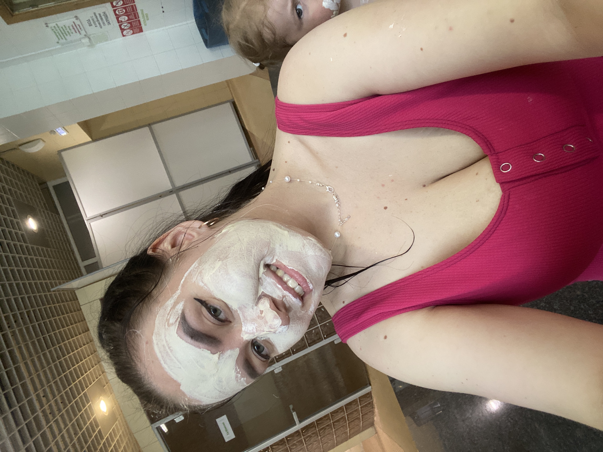 A woman with a white facial mask smiles at the camera. She is wearing a pink top and a delicate necklace. A child is partially visible in the background, also with white cream on their face. The location appears to be indoors, possibly a bathroom or spa, given the tiling, fixtures, and signage visible in the background. The city is Votkinsk, Russia.