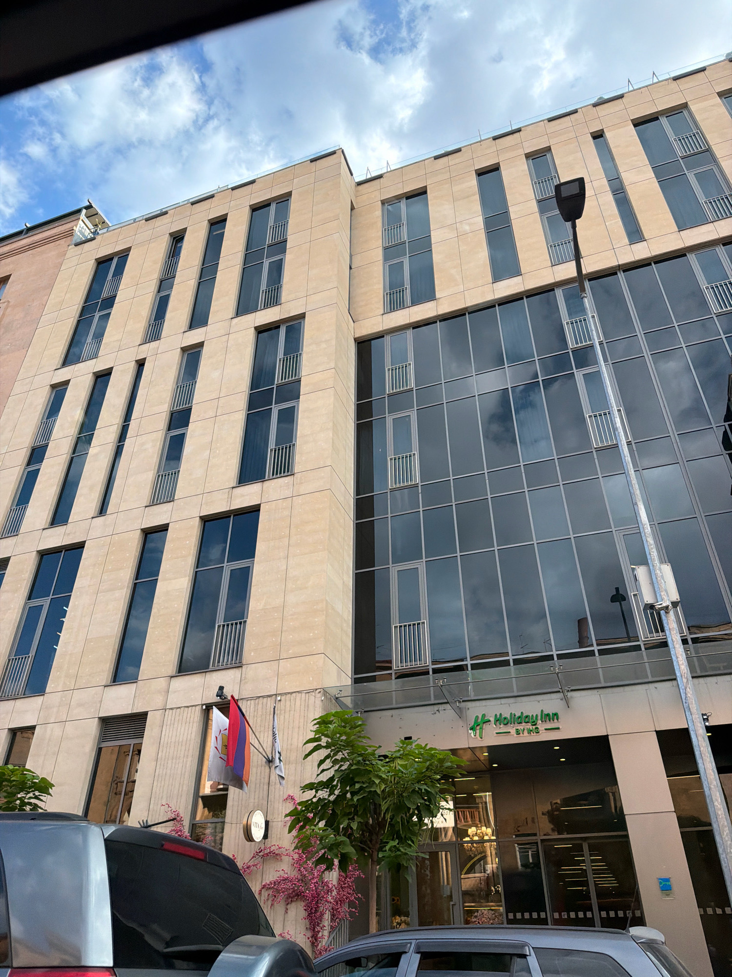 A medium shot captures the exterior of a modern, multi-story building in Yerevan, Armenia, under a partly cloudy sky. The building features a facade of light-colored stone cladding interspersed with dark, reflective glass windows, many of which have small, barred balconies. A street lamp stands prominently in the foreground, slightly to the right of center.
On the building's facade, a "Holiday Inn BY IHG" sign is visible, indicating the presence of a hotel. Two flags, one of which appears to be the Armenian flag, are mounted on the building near the entrance. Lush green trees and vibrant pink flowering vines add a touch of nature to the urban landscape. In the lower portion of the frame, the rear of a silver SUV is visible, partially obscuring the building's lower levels and entrance. The overall impression is one of a contemporary urban setting.