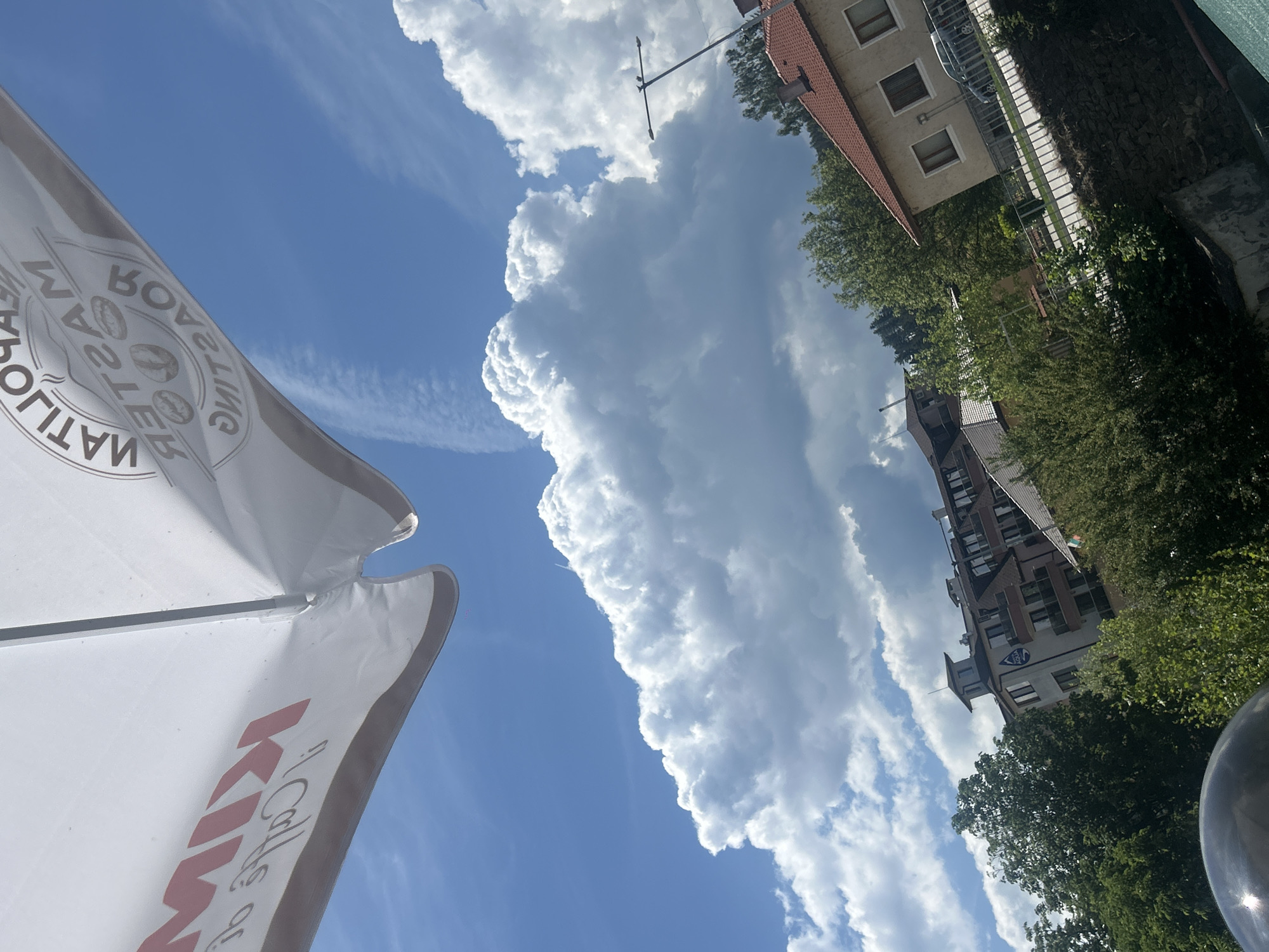 The image captures an outdoor scene under a bright blue sky dotted with large, fluffy cumulus clouds and some wispy cirrus clouds, indicating midday or early afternoon fair weather. In the foreground, partially visible on the left and bottom, is a large white patio umbrella. Its surface bears mirrored text: "MATILBOV" (reversed) on the upper left section, and a red "KIMBO Caffè" logo on the lower right section, indicating a coffee brand.
Beyond the umbrella and beneath the expansive sky, an urban or semi-urban landscape unfolds. On the right side of the frame, several buildings are visible amidst lush green trees and bushes. Some buildings feature distinctive red tile roofs, common in the region, while others are taller and more modern, with multiple windows and balconies. One of these modern buildings, located in the mid-right, displays a blue logo with the word "AQUA" on its facade. A street light or utility pole extends into the sky near the center-right. A metallic, spherical object, possibly part of another lamp or a reflective surface, is partially seen in the far bottom right. The overall impression is one of a pleasant, calm summer day in Bania, Bulgaria, likely taken from a seating area outside a cafe.