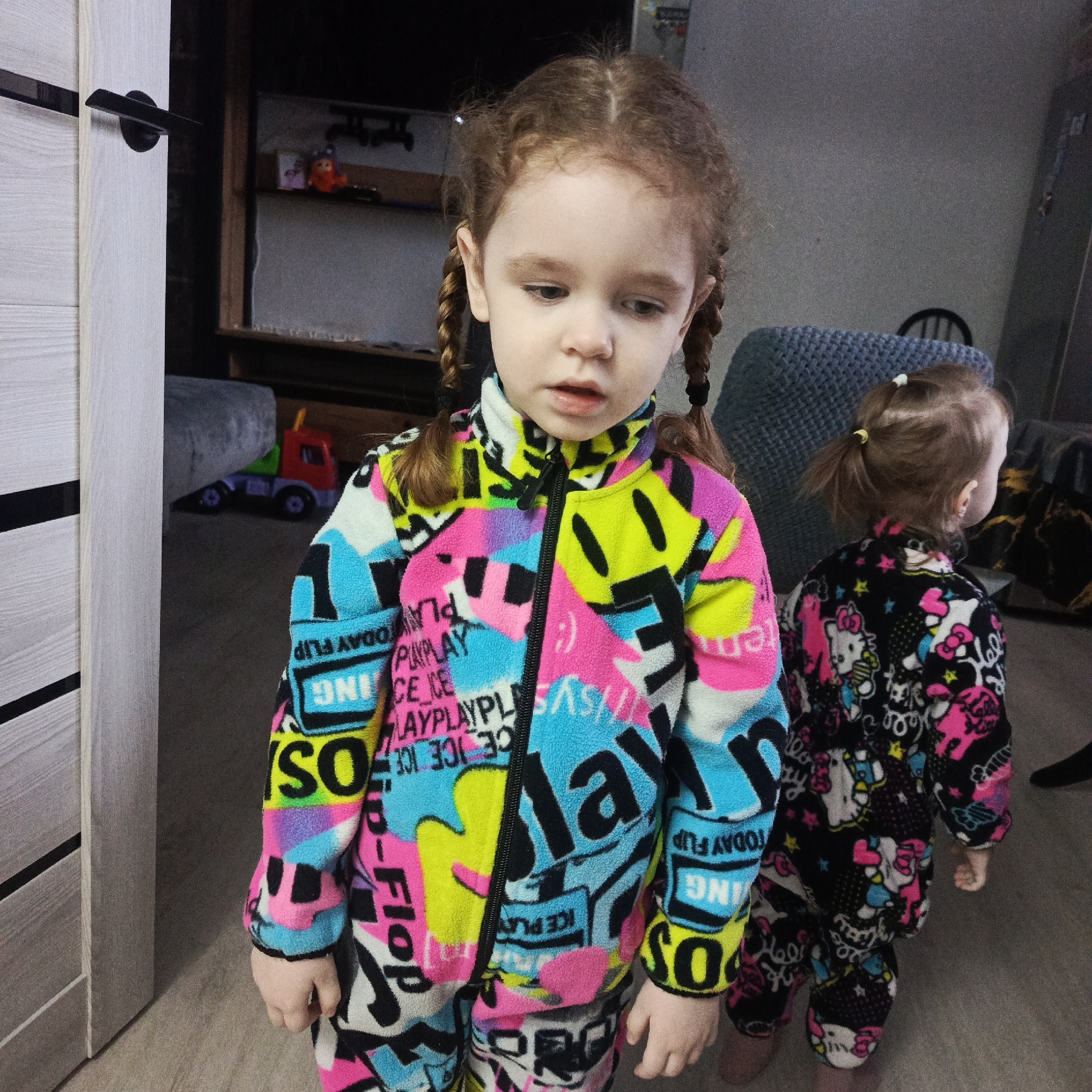 The image shows two young girls in an interior space. The girl in the foreground has light skin, red hair styled in braids, and wears a brightly colored fleece jumpsuit with a chaotic pattern of words and shapes in pink, yellow, blue, black, and white. The jumpsuit is zipped up to the neck. She is looking down. In the background, a second girl, slightly smaller, has her hair in a ponytail and wears a dark jumpsuit patterned with pink and white Hello Kitty images. She faces away from the camera.
In the background is a grey chair and a unit with shelves, on which sit some toys. A portion of a closed white door is visible on the left. The floor appears to be light wood. The image location is in the city of Марковский, Russia.