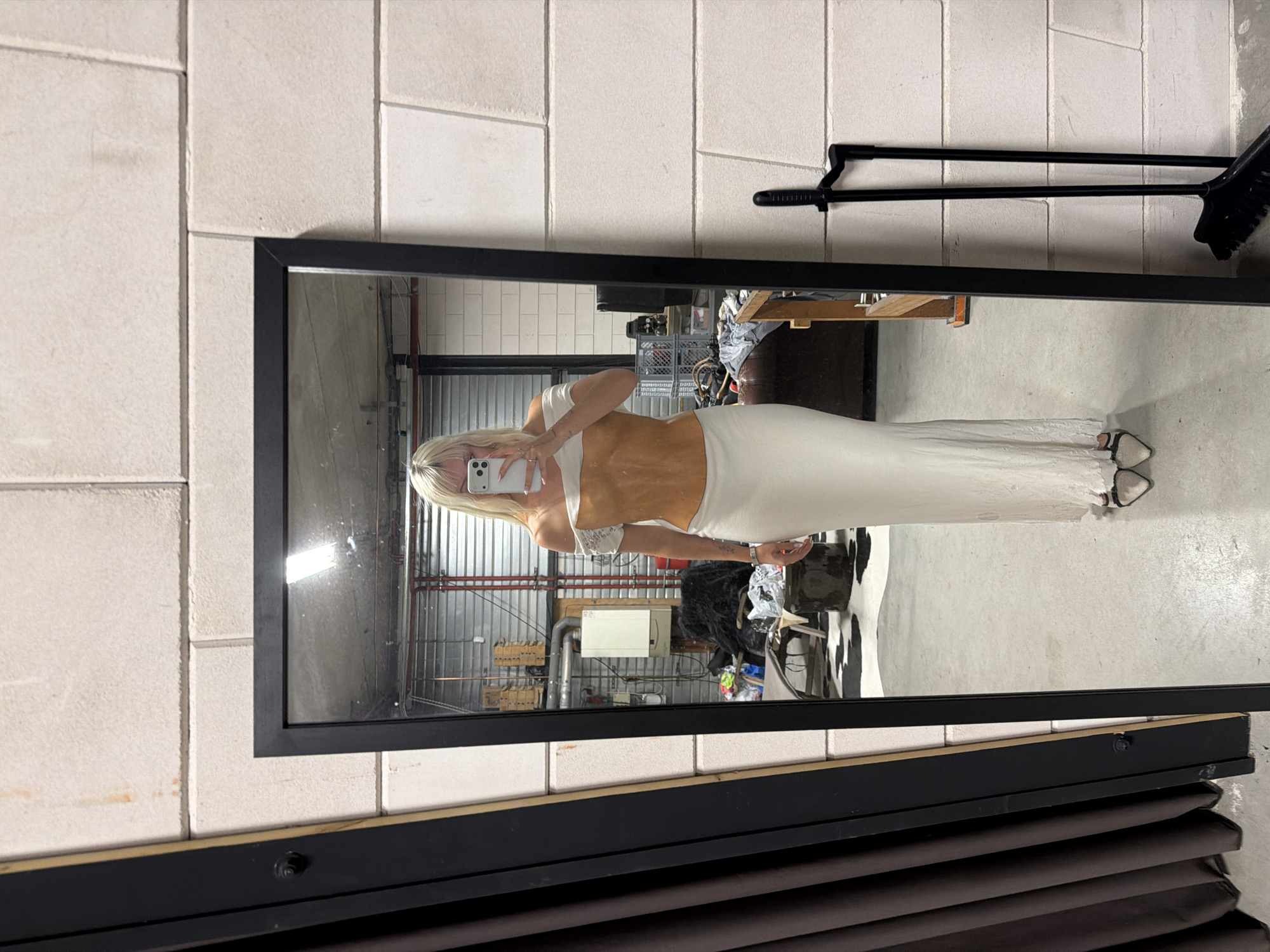 A woman with blonde hair stands in front of a full-length mirror, taking a selfie with her phone. She is wearing a white off-the-shoulder top and a long white skirt. The mirror reflects a room with exposed brick walls, industrial-style shelving, and various objects on the floor. A broom and dustpan set stands to the right of the mirror. The scene appears to be an indoor space, possibly a workshop or studio, not a location in Amsterdam, Netherlands.