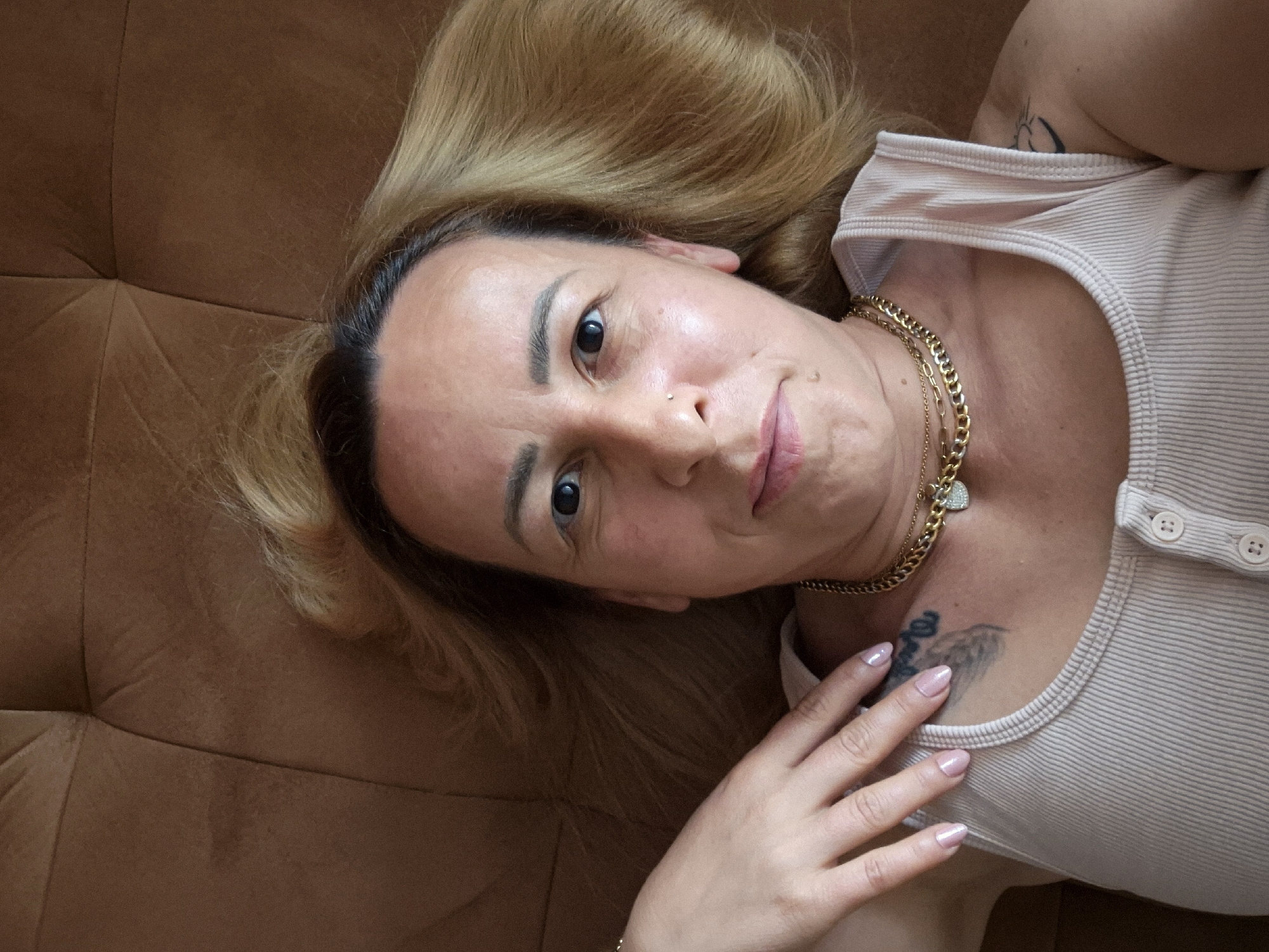 In this overhead, close-up, selfie-style image, a woman with blonde hair lies on a brown textured surface, possibly a couch. She is wearing a light-colored, ribbed tank top with a button detail at the neckline and several layered gold necklaces, one with a heart-shaped pendant. Her left hand, with light pink nail polish, rests on her shoulder. A tattoo is visible on her left shoulder and another on her right shoulder. She has a nose ring and appears to be looking at the camera with a slight smile. The background is a solid brown color with visible stitching or texture. The lighting is bright, creating some shine on her skin.