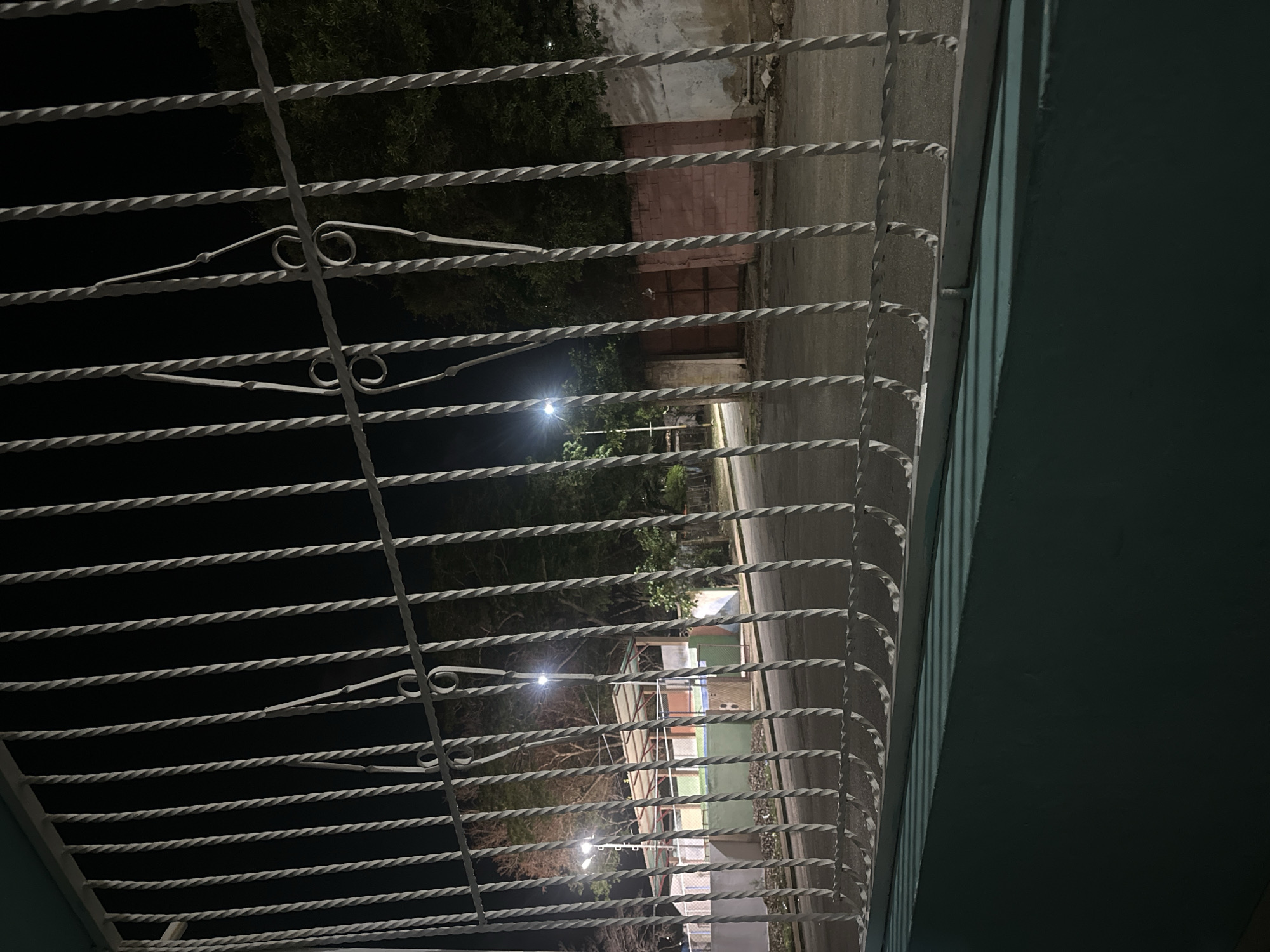 The image shows a view through a white metal gate at night. Beyond the gate, a street is visible with a sidewalk and a building with a covered outdoor area. There are streetlights casting illumination on the scene. Trees are also present in the background. The foreground consists of the lower part of a teal-colored wall and the base of the white gate. No people are visible in the image. The scene appears to be in a residential or recreational area.