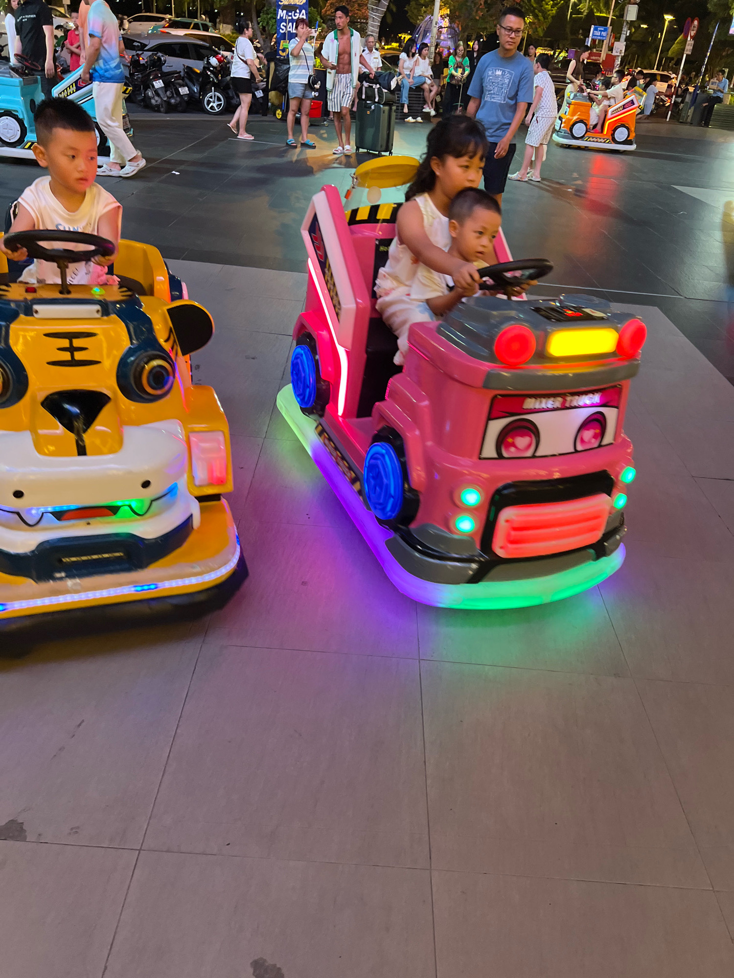 In Nha Trang, Vietnam, two children are riding in motorized toy vehicles on a tiled pedestrian area, likely a promenade or entertainment zone. The child in the foreground is a young boy in a white t-shirt, seated in a yellow and blue toy vehicle designed to resemble a tiger. He is holding a black steering wheel. To his right, a pink toy truck is occupied by a young girl wearing a light-colored outfit, and a younger boy, who is also holding a steering wheel. The toy vehicles are illuminated with colorful LED lights. In the background, other people and vehicles are visible, suggesting a public space with ongoing activity, possibly at dusk or evening given the lighting. The overall impression is one of a recreational setting in a city.