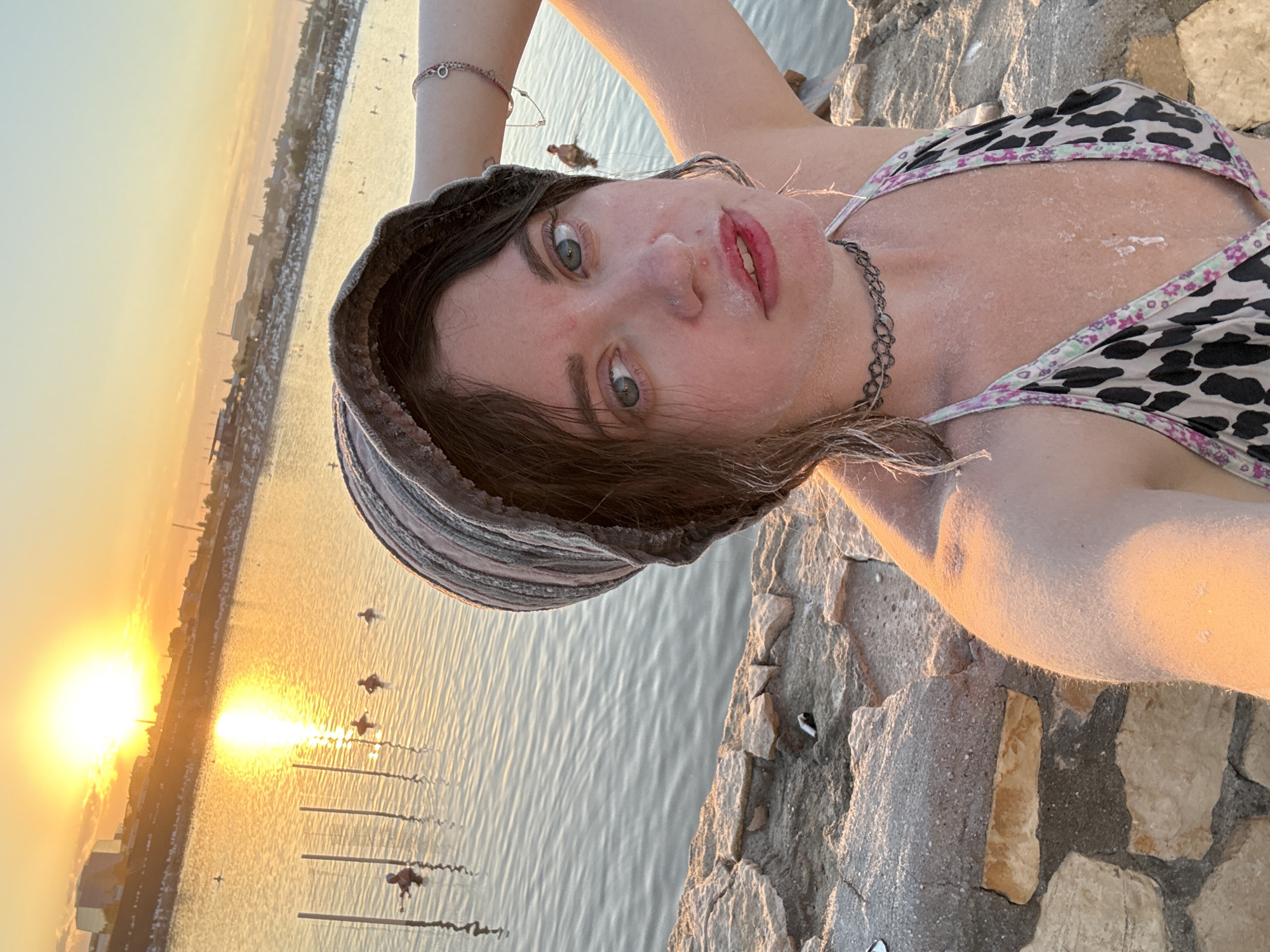 This is a selfie taken by a young woman in the city of Sol-Iletsk, Russia. The woman is wearing a bucket hat and a bikini top, and her skin is covered in salt crystals, indicating she is at a salt lake or similar environment. The sun is setting, casting a warm golden light over the scene, with the sun prominently visible in the upper left quadrant of the image. The background shows a body of water, with several other people visible swimming or wading. Buildings and a shoreline are also present in the distance. The woman is positioned in the foreground, with a stone embankment behind her.