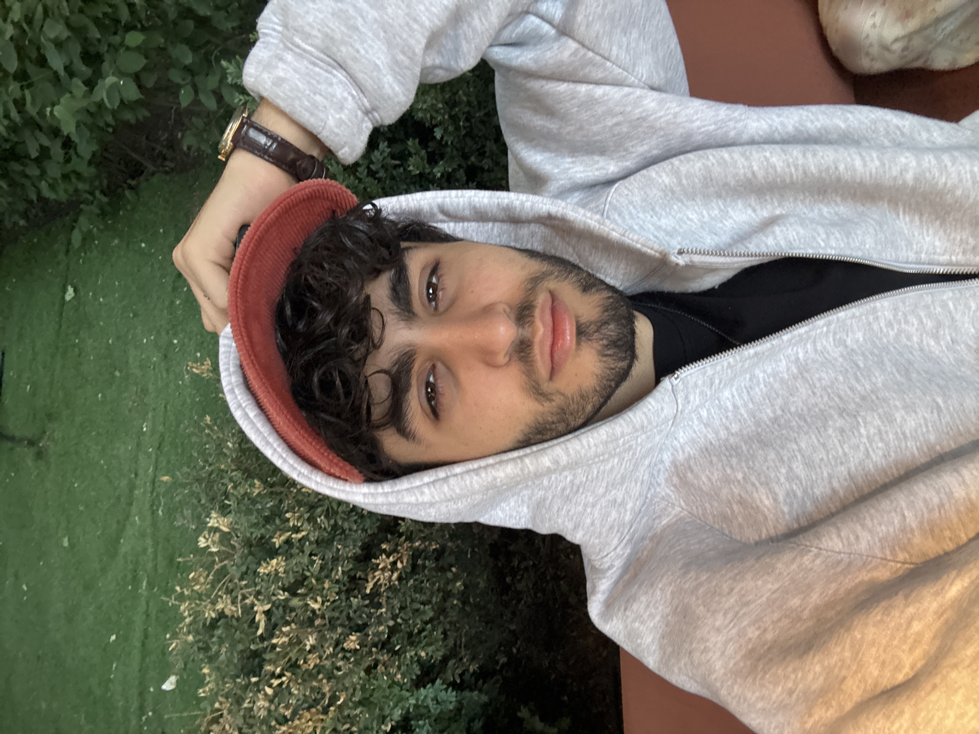 The image, originally rotated 90 degrees counter-clockwise, displays a close-up portrait of a young man, likely a selfie. He has dark, curly hair and a well-groomed beard or prominent stubble. His eyes are dark brown, and his lips are full, appearing slightly pursed.
The man is wearing a light grey hooded sweatshirt with a zipper, under which a black t-shirt is visible. On his left wrist, he sports a classic watch with a gold-toned case and a dark brown leather strap. He is holding an orange-red cap, possibly made of corduroy, by its brim, positioning it near his head.
The background shows lush green vegetation, primarily dense bushes or hedges, and a patch of green grass, suggesting an outdoor environment such as a garden or park. To the right of the man, a reddish-brown surface, possibly a table or bench, is visible, against which he may be leaning or resting.
The lighting appears soft and natural, indicating daytime, possibly late afternoon. The man is looking directly into the camera with a neutral to slightly pensive expression. There is no visible text in the image. The setting is consistent with an outdoor location in Yerevan, Armenia, as specified.
