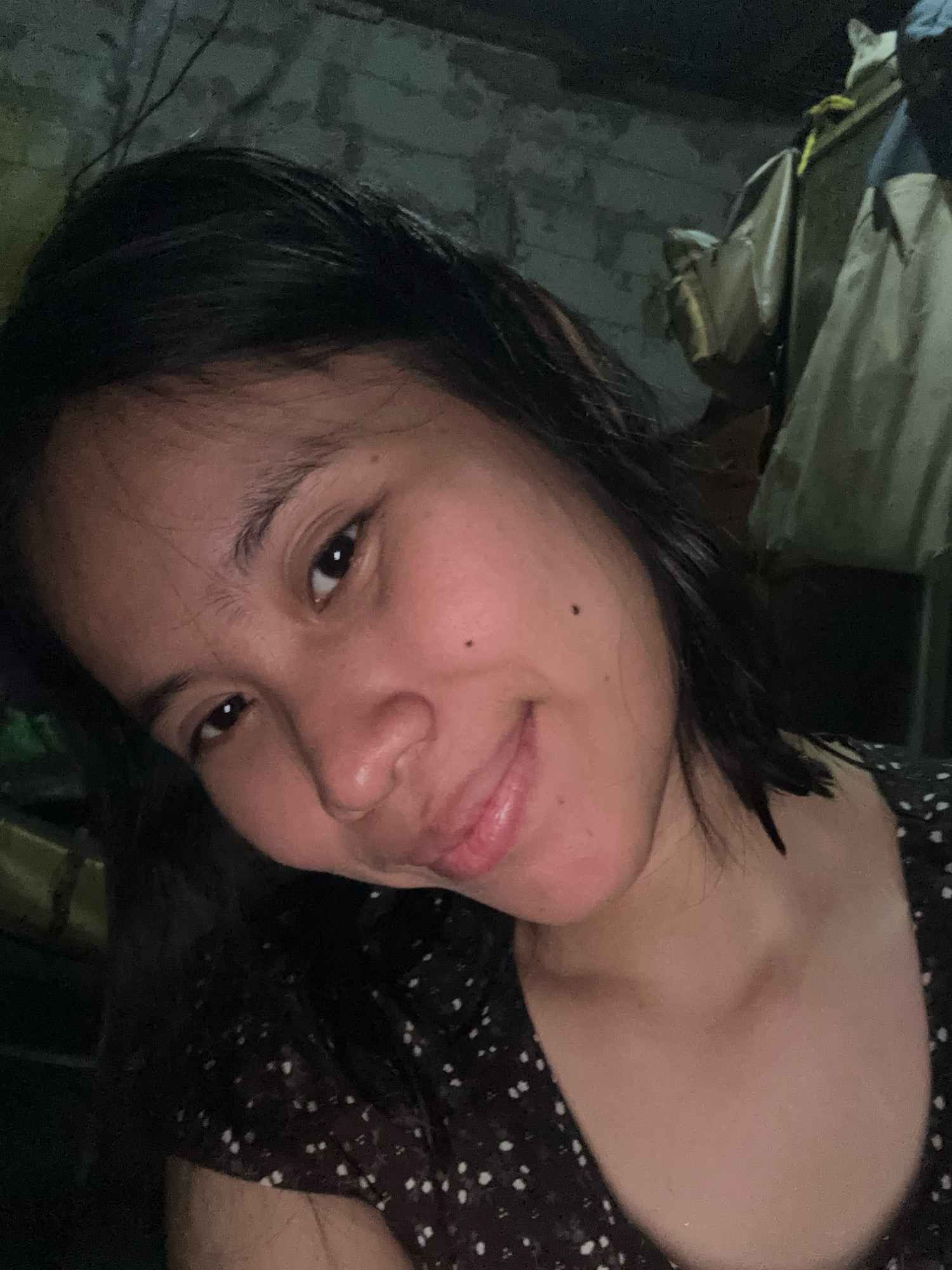 In a dimly lit, indoor environment, a young woman with dark hair and a brown polka dot top poses for a selfie. Her head is tilted, and she is looking towards the camera with a slight smile and her tongue slightly out. The background appears to be a textured wall with some objects hanging on it, possibly clothing or bags. The overall scene suggests a casual, personal moment captured in East Jakarta, Indonesia.