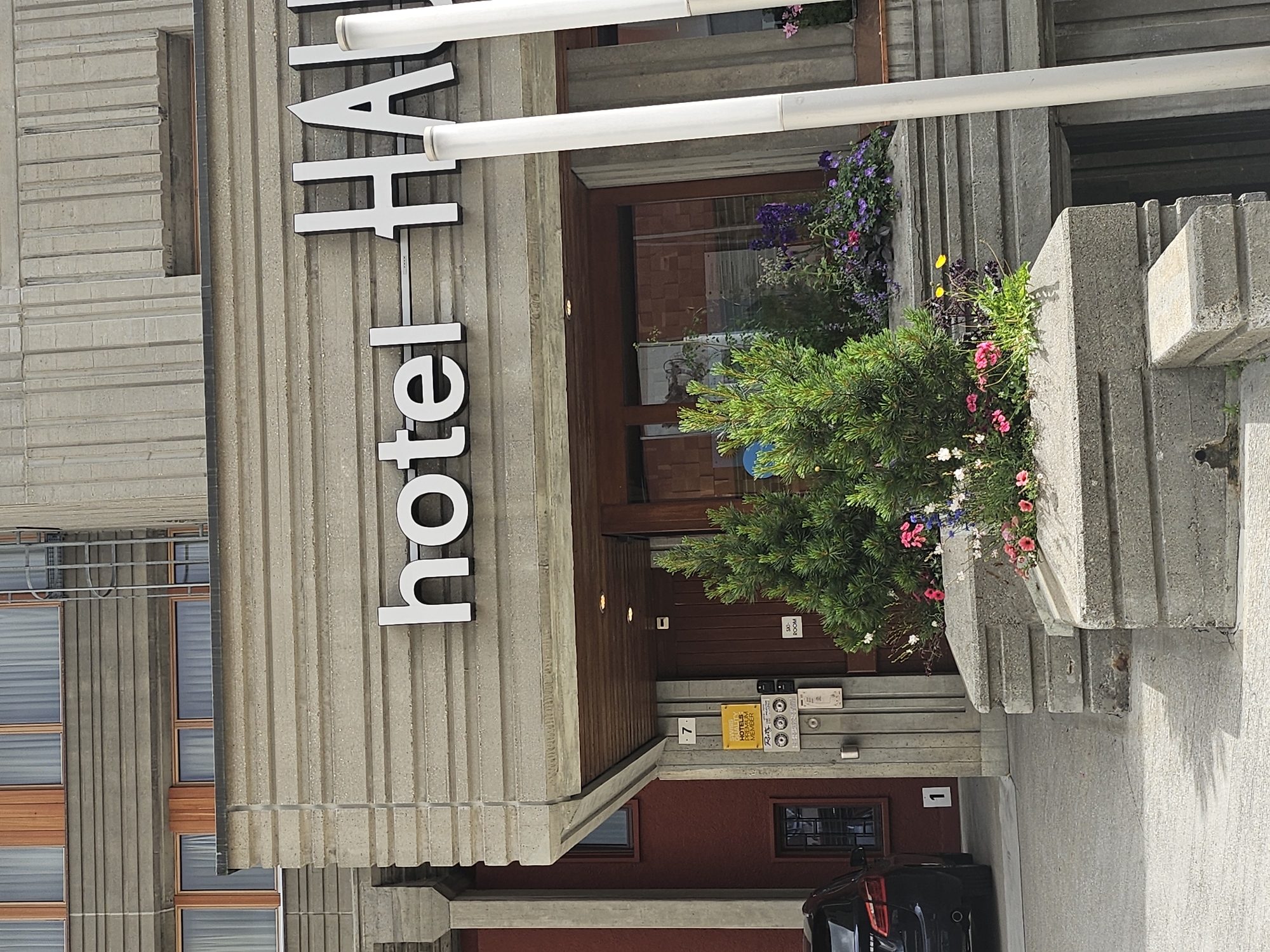 The image shows the exterior of the Hotel HAUS post-fire in St. Moritz, Switzerland, with a sign above the entrance reading "hotel HAUS". The hotel's facade is made of concrete with horizontal grooved detailing. To the left of the entrance, a dark-colored car is parked, and the number "1" is visible on a wall. Adjacent to the entrance, a yellow sign indicates the hotel is a "SMARTY HOTELS MEMBER". A small green Christmas tree is planted in front of the entrance, surrounded by various flowering plants. The entrance itself features a wooden door and is under a covered walkway. The weather appears to be fair.
