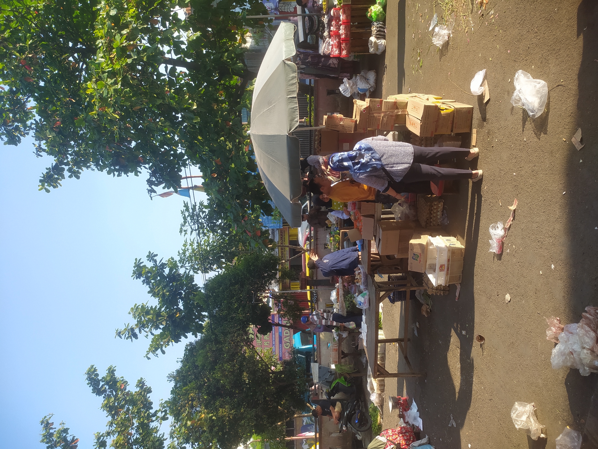 This image depicts an outdoor market scene under a clear blue sky, likely in Kupang, Indonesia, given the context provided. The market is bustling with activity, featuring several people engaged in transactions or arranging goods. Various stalls are set up, displaying a range of products, including what appear to be vegetables, eggs, and packaged items. A large gray umbrella provides shade over one of the stalls. The ground is paved and shows signs of recent activity, with some discarded plastic bags and debris scattered around. The presence of trees, some with green foliage and others with hints of red leaves, suggests a tropical or subtropical climate. In the background, buildings and vehicles, including a blue truck, indicate an urban setting. The overall impression is one of a lively and active marketplace during daylight hours.