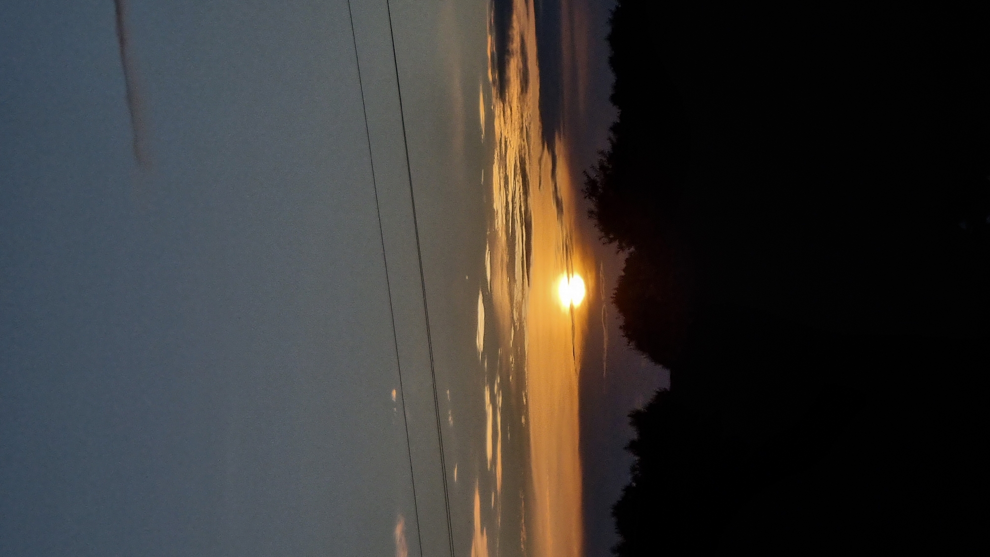 A vertical, full-frame shot captures a sunset over a silhouetted treeline in the city of Vysokoe, Russia. The sun, a bright orb of orange-yellow, is positioned slightly left of the center of the frame, just above the horizon. It is partially obscured by a layer of dark clouds. Above the sun, thin horizontal lines of power lines stretch across the sky. The sky is a gradient of deep blue at the top, transitioning to lighter shades of blue and orange near the horizon, with scattered, wispy clouds. The foreground is entirely black, with the shapes of trees and possibly some low-lying structures visible as dark silhouettes against the brighter sky. The image has a slightly grainy texture, consistent with low-light photography.