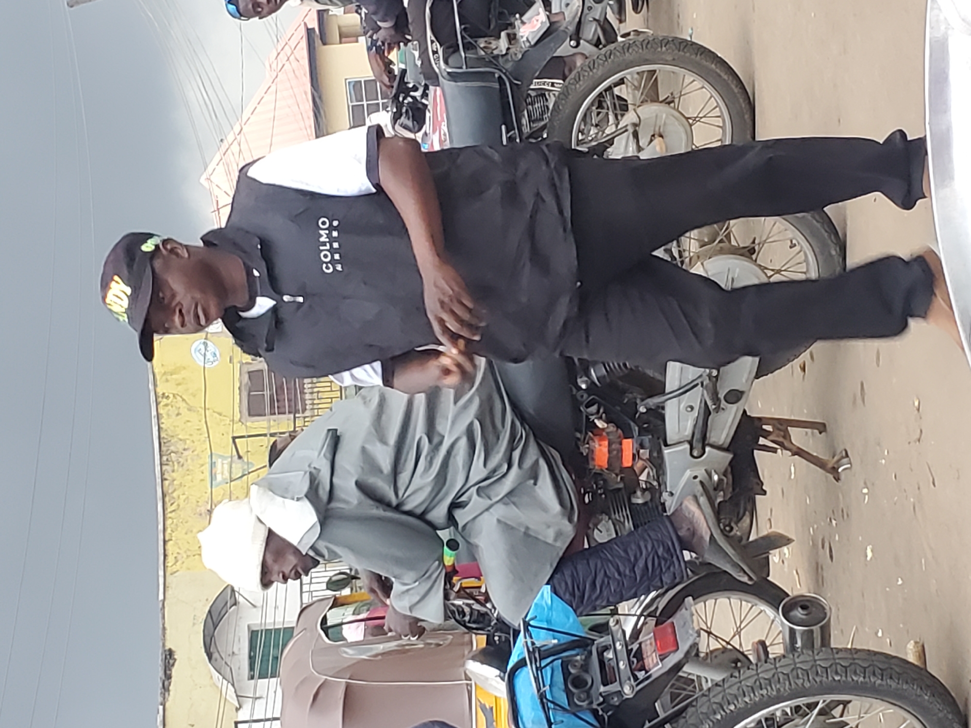 In Gwong, Nigeria, two men are seen on motorcycles. The man in the foreground, wearing a black vest with "COLMO" printed on it and a black cap, stands next to a motorcycle. His hands are visible as he holds what appears to be a small object. Behind him, another man, dressed in a grey raincoat and white head covering, is seated on a motorcycle. He is looking to his left. The background shows buildings and overcast skies, typical of a street scene in the city.
