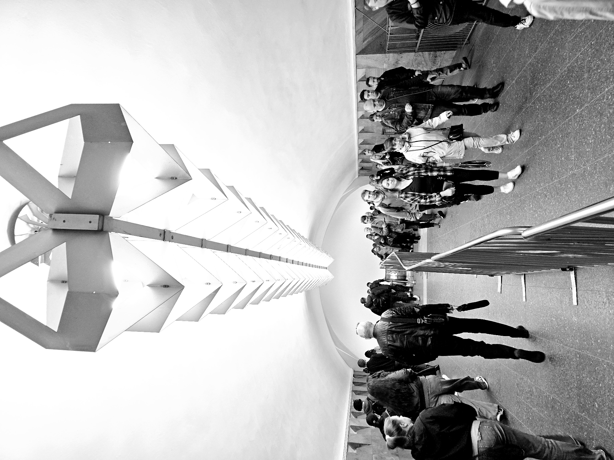 The image depicts a black and white photograph taken inside a metro station in Moscow, Russia. A long row of people are waiting in line behind a metal barrier. An overhead light fixture consists of geometric shapes resembling a spine extending into the distance. The walls of the station are curved, converging in the background.