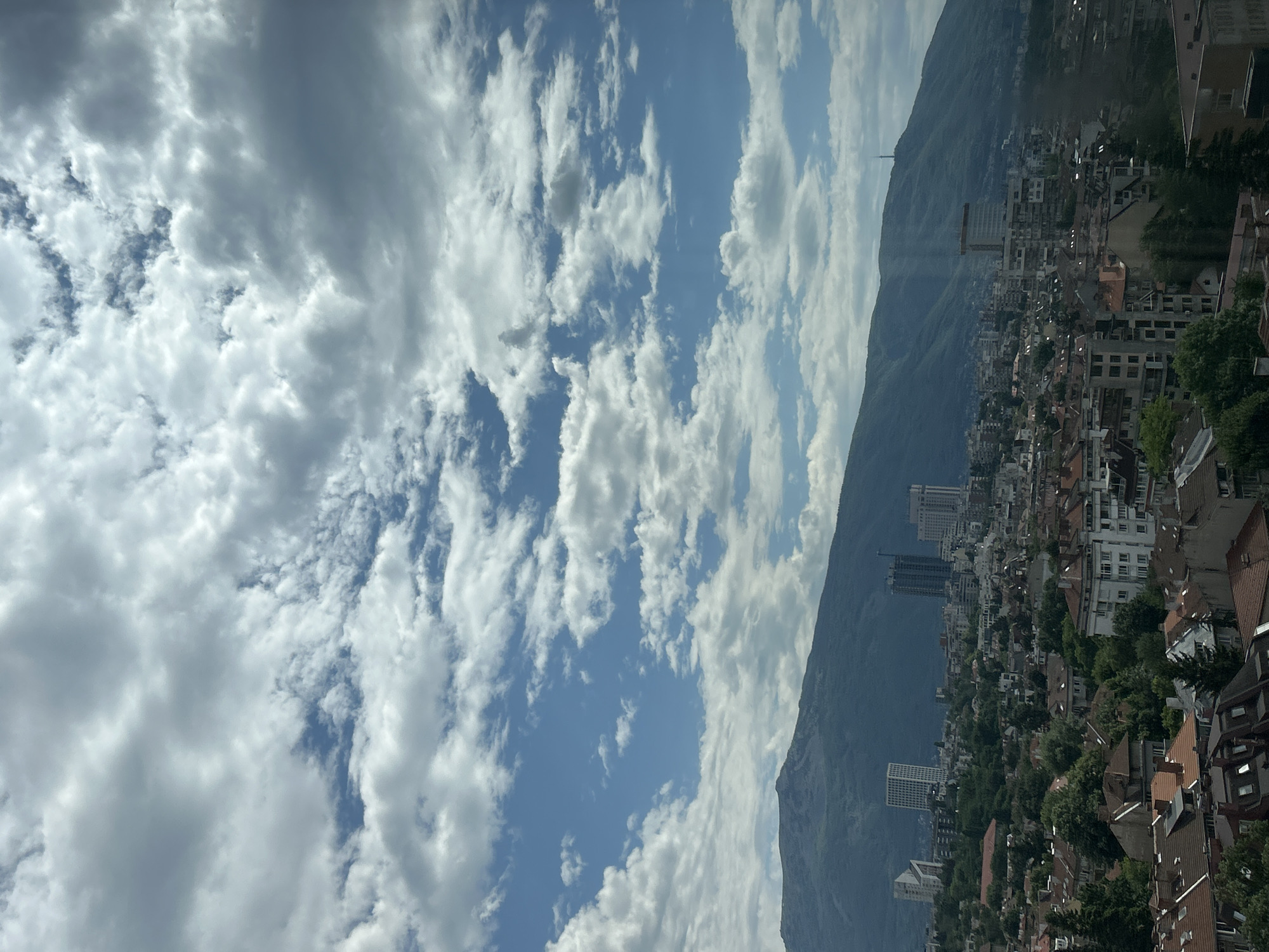 The image presents a panoramic daytime view of a city nestled against a majestic mountain range under a partly cloudy sky. The photograph is originally oriented vertically, rotated 90 degrees counter-clockwise, meaning the mountain forms the lower horizon and the sky is above.
The upper portion of the corrected image is dominated by a vibrant blue sky dotted with numerous cumulus clouds of varying sizes and densities. Some clouds are bright white and fluffy, while others appear larger and a deeper grey, suggesting depth and movement in the atmosphere.
Below the sky, the imposing dark green and grey mass of a mountain stretches across the horizon, serving as a dramatic natural backdrop to the urban sprawl. Near one of the peaks on the mountain, a slender, dark structure, likely a communications tower or antenna, is visible. Shafts of sunlight appear to break through the clouds, casting subtle rays of light across the mountain slopes and parts of the city.
The lower half of the image showcases a sprawling cityscape. A dense cluster of buildings, primarily residential, fills the foreground and middle ground. The architecture features a mix of multi-story buildings with light-colored facades and numerous roofs sporting shades of red, orange, and brown tiles. Interspersed among these are several taller, more modern structures. Green trees are visible throughout the city, breaking up the urban landscape and indicating parks or tree-lined streets.
The overall setting suggests a pleasant day with good visibility. Given the context provided, this is a view of Sofia, Bulgaria, with the prominent Vitosha Mountain in the background. No people or visible text are discernible in the image.
