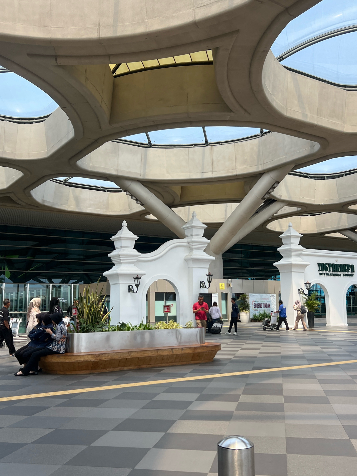The image captures the exterior of Yogyakarta International Airport in Palihan, Indonesia. The architecture features a distinctive, undulating concrete roof structure with large, open circular cutouts that allow natural light to filter through, revealing the sky above. Supporting the roof are substantial concrete pillars. In the foreground, a checkered grey and black tiled pedestrian area is visible, with a yellow tactile paving line running across it. A modern, wooden bench with a metallic planter box is situated in the mid-ground, containing some green foliage.
Several people are present, engaged in various activities. On the left, a group of individuals, some wearing hijabs, are sitting on the bench. Further to the right, people are walking and pulling luggage, indicating travel. One person in a red shirt is looking at their phone, while others appear to be moving towards or away from the airport entrance. The entrance itself is marked by ornate white pillars and arched gateways, with signage that includes the text "YOGYAKARTA INTERNATIONAL AIRPORT" and "SUGENG TINDAK". The building's facade is glass-fronted, reflecting the sky and surroundings. The overall atmosphere is one of a busy transportation hub on a clear day.
