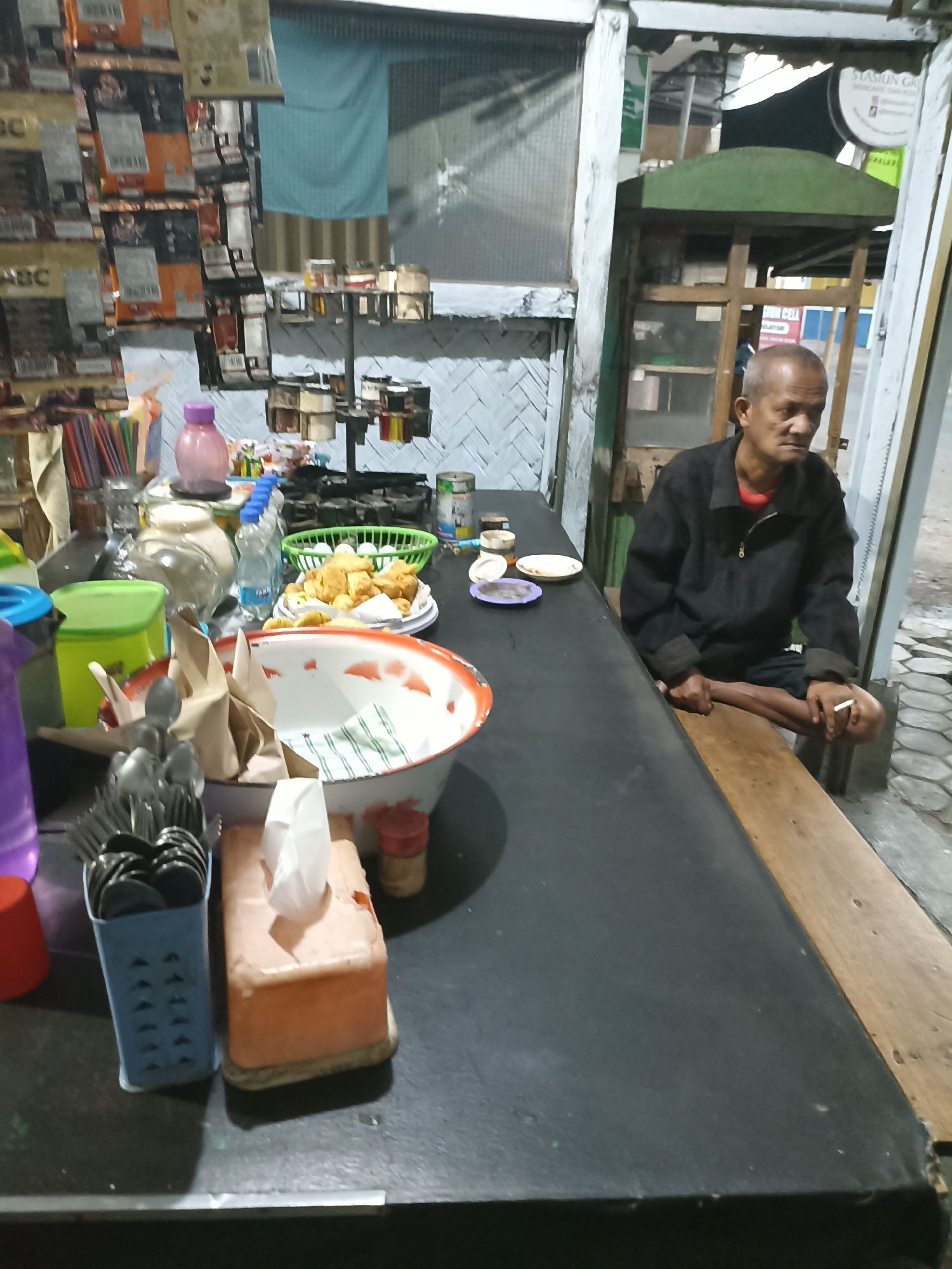 The image depicts a street-side food stall, likely in Lumajang, Indonesia. An elderly man with a shaved head, wearing a black zip-up jacket over a red shirt and dark shorts, is seated on a wooden bench. He is smoking a cigarette, holding it in his right hand. The stall counter is black and cluttered with various items. In the foreground, there is a white enamel bowl with red trim, containing folded napkins. Next to it is a tissue box with a white tissue protruding. To the left of the bowl, a blue container holds silverware. Behind the bowl, a green basket holds fried food items and eggs. Other items visible on the counter include bottles of water, jars, and containers of spices. The stall's background features shelves stocked with packaged goods, and a blue tarp hangs behind them. The overall setting appears to be an outdoor area, with a tiled ground visible to the right.