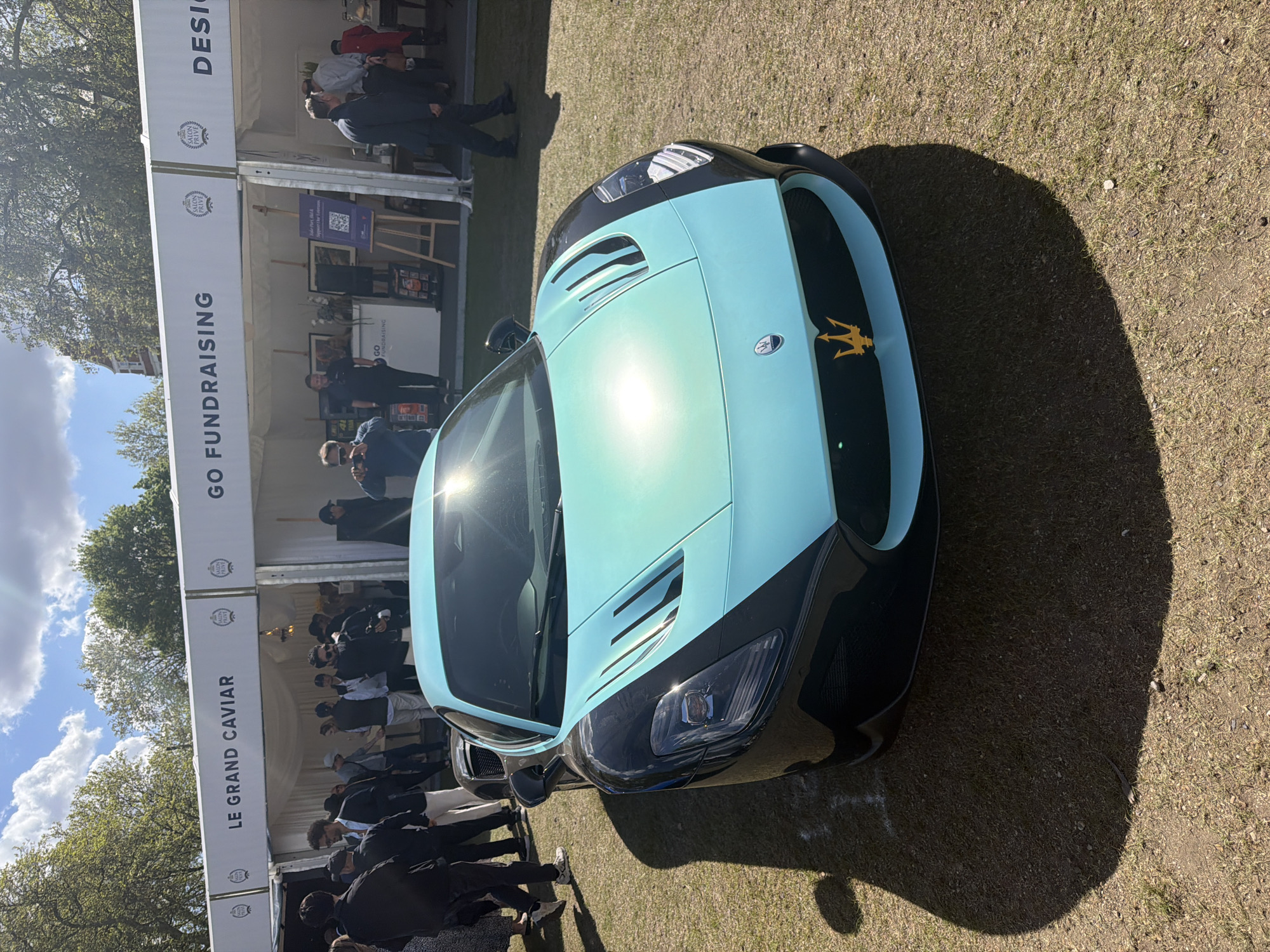 A vibrant light blue and black Maserati MC20 sports car is prominently displayed outdoors on a sunny day. The car, positioned on a grassy patch, features a distinctive light blue body contrasting with black accents, including parts of the front bumper and grille. A golden Maserati trident emblem is clearly visible on the front. Bright sunlight reflects off the car's hood and windshield.
In the background, two large white marquees or event tents are set up, hosting numerous people. Attendees are seen milling around and conversing, suggesting an ongoing event or exhibition. The ground is dry grass with some patches of dirt. Trees with green foliage and a partly cloudy blue sky are visible above the tents.
Notable details include long shadows cast by the car and people, indicating a bright daytime setting, likely morning or early afternoon. Visible text on the tents includes "LE GRAND CAVIAR", "GO FUNDRAISING", and multiple instances of "SALON PRIVE", along with a partially visible "DESI". These elements suggest an upscale fundraising event or car show, possibly part of the "Salon Privé" event series, located in London, United Kingdom. The overall atmosphere appears lively and celebratory.