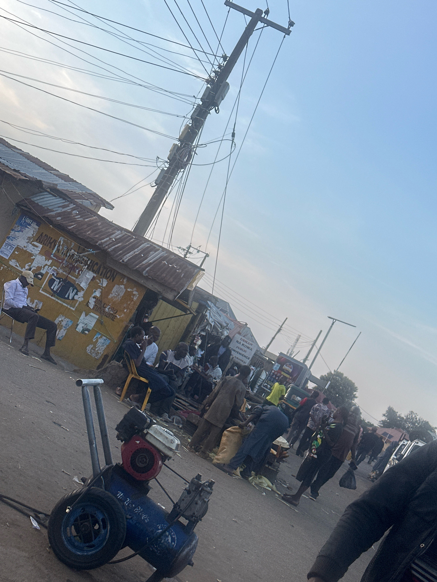 The image depicts an outdoor market scene in Gura Top, Nigeria, under a hazy sky with visible power lines overhead. In the foreground, a blue air compressor with a red engine sits on the asphalt road. To the left, a yellow structure with a corrugated metal roof appears to be a small shop or kiosk, adorned with posters and signs, including one that reads "ARIKAN LIKION EDUCATION" and another with "MTN". A man in a white shirt and beige cap sits on a white chair outside this structure, looking at his phone.
Further into the scene, a crowd of people are gathered, some sitting on yellow chairs and others standing. Several individuals are engaged in conversation or other activities. In the background, more stalls and structures are visible, along with trees and more power lines. A green tricycle is also present, suggesting transportation in the area. The overall atmosphere appears to be that of a busy marketplace.