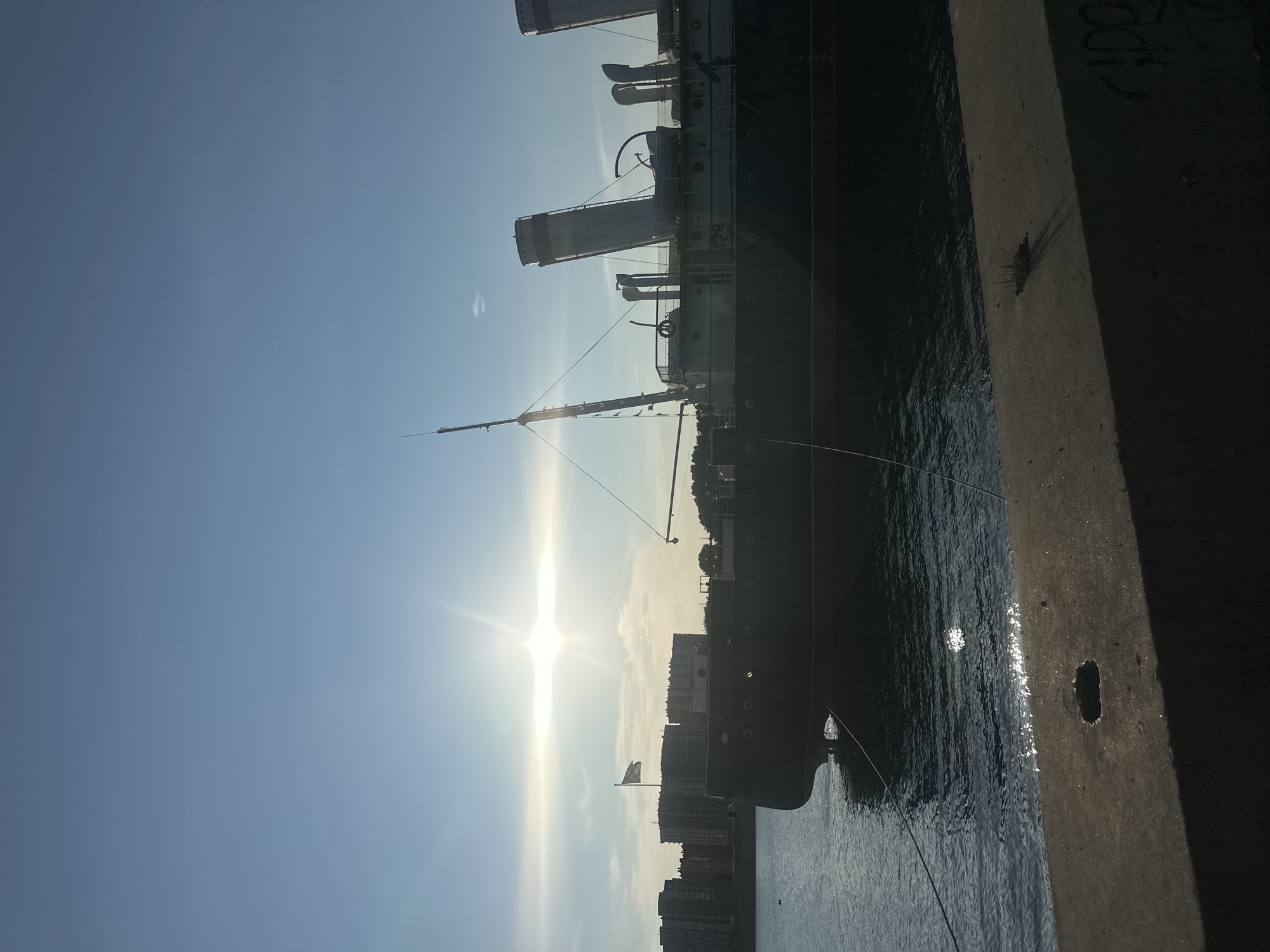 This media file is a vertical, slightly tilted photograph taken outdoors. The primary subject is the side of a large ship, likely a museum ship, docked next to a concrete pier. The ship appears old, with multiple decks, smokestacks, and rigging visible. The pier is rough and weathered, with graffiti visible in the upper right portion of the frame.
The scene is set by a bright, low sun, indicating it is either early morning or late afternoon. The sky is a clear, pale blue with some wispy clouds. The sun's rays are prominent, creating a lens flare effect and reflecting off the calm water surrounding the ship. In the distance, behind the ship, is a line of modern buildings, suggesting an urban waterfront location. A flag is visible on a pole near the ship.
No people are visible in the image. The overall mood is one of quiet observation, with the strong sunlight and clear sky suggesting a pleasant day. The graffiti on the pier, including what appears to be "AIDOV" is a notable detail. The location context of Irkutsk, Russia, is provided externally.