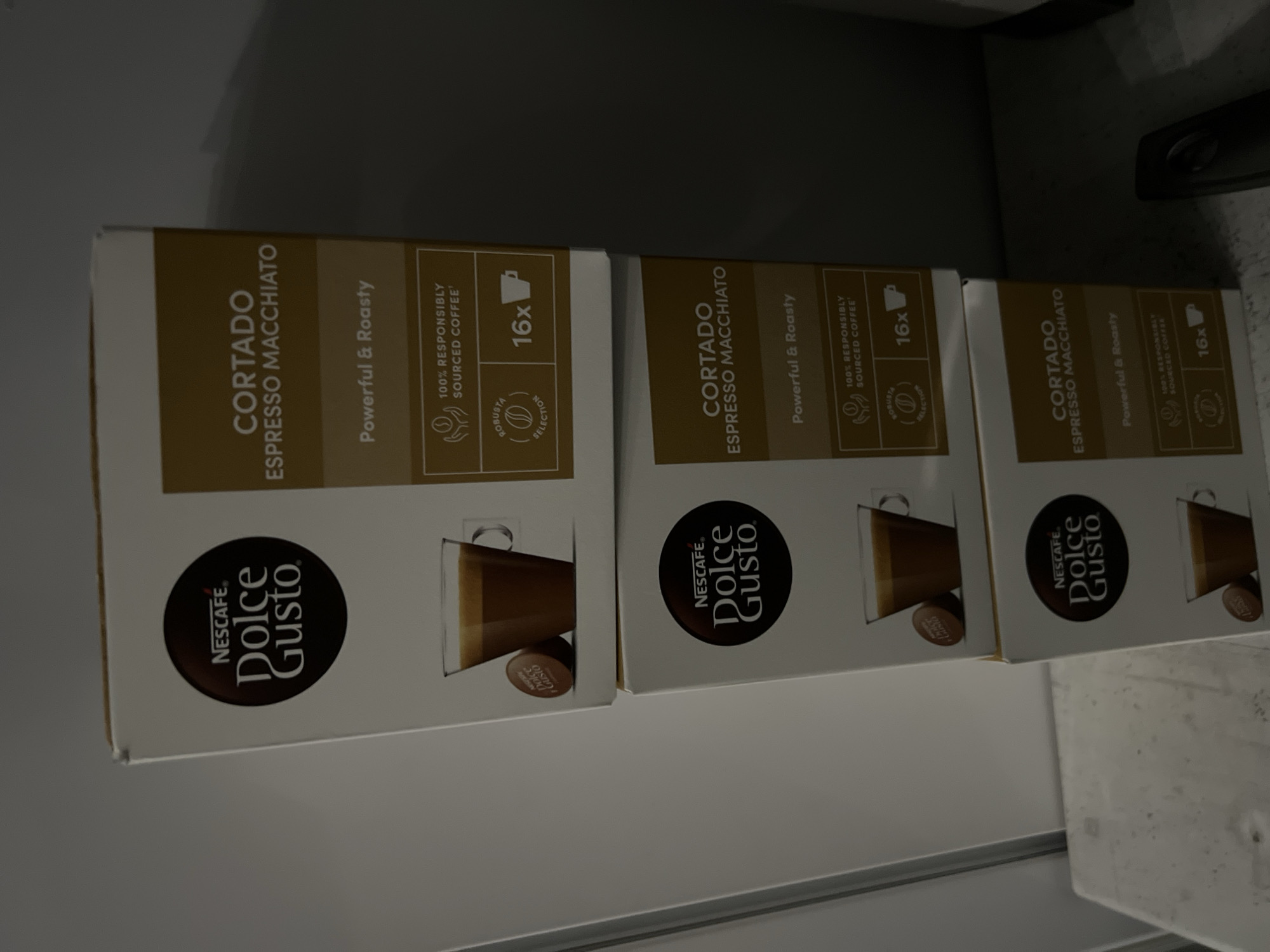 Three identical boxes of Nescafé Dolce Gusto Cortado Espresso Macchiato coffee capsules are displayed in an indoor setting in Burgas, Bulgaria. The boxes are arranged in a slight diagonal line on a flat, light-colored surface, possibly a shelf or counter, with a plain, darker background. The lighting is soft and ambient.
Each box is primarily brown and white, featuring the circular "NESCAFÉ. Dolce Gusto." logo. The product name "CORTADO ESPRESSO MACCHIATO" is printed vertically on the front. Below this, descriptors such as "Powerful & Roasty" are visible. Further details indicate "100% RESPONSIBLY SOURCED COFFEE" and highlight a "ROBUSTA SELECTION." Each package contains "16x" capsules, as denoted by a number next to a coffee cup icon. A graphic on the lower portion of each box illustrates a glass of Cortado espresso macchiato and a single coffee capsule. The boxes appear new and unopened, suggesting they are either fresh stock or recently acquired.