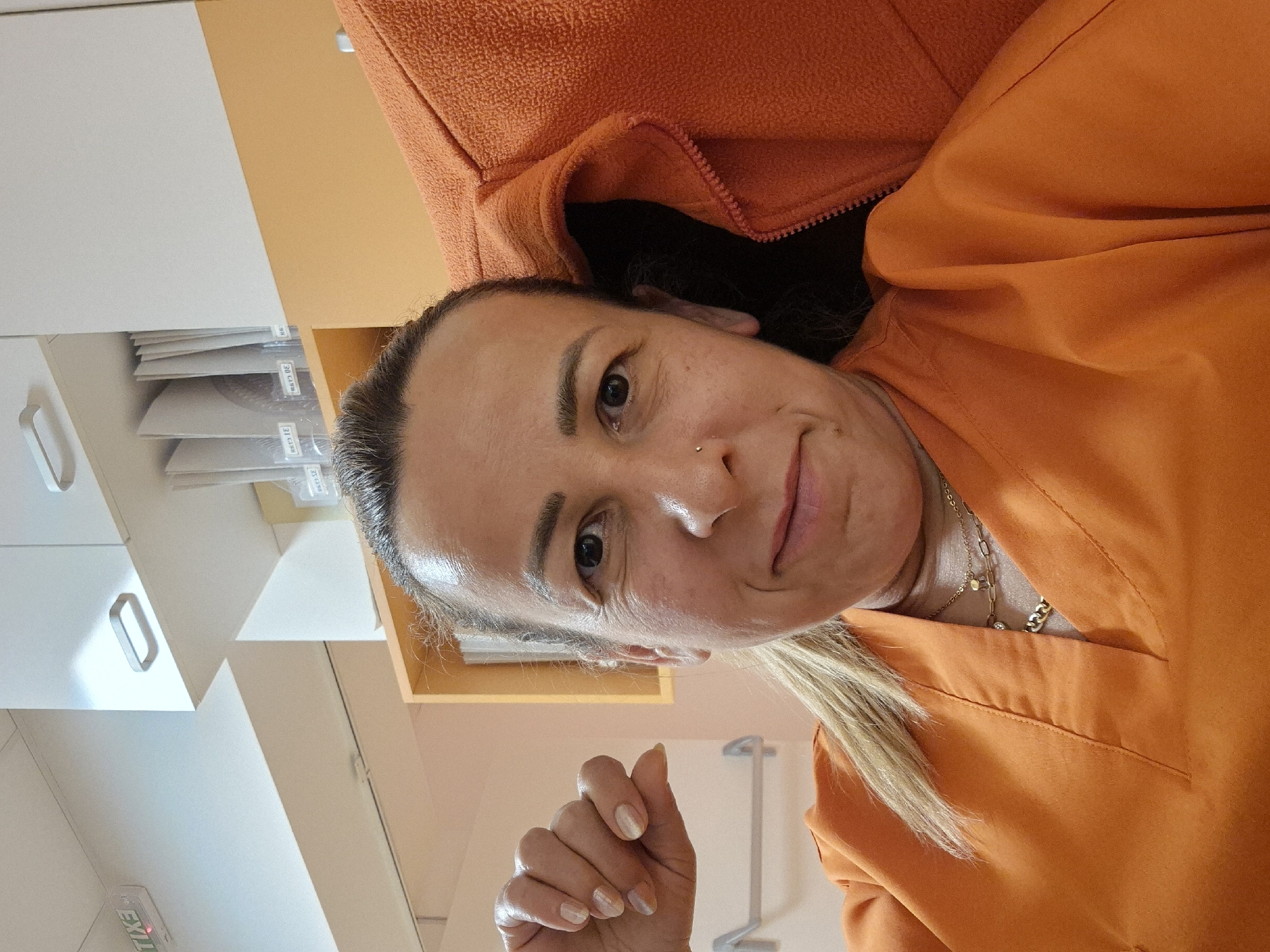 A woman is taking a selfie in an office. She is wearing an orange scrub top and a light brown jacket. She has blonde hair pulled back in a ponytail and is wearing jewelry. In the background, there are cabinets, shelves, and an exit sign. The room appears to be well-lit and tidy.