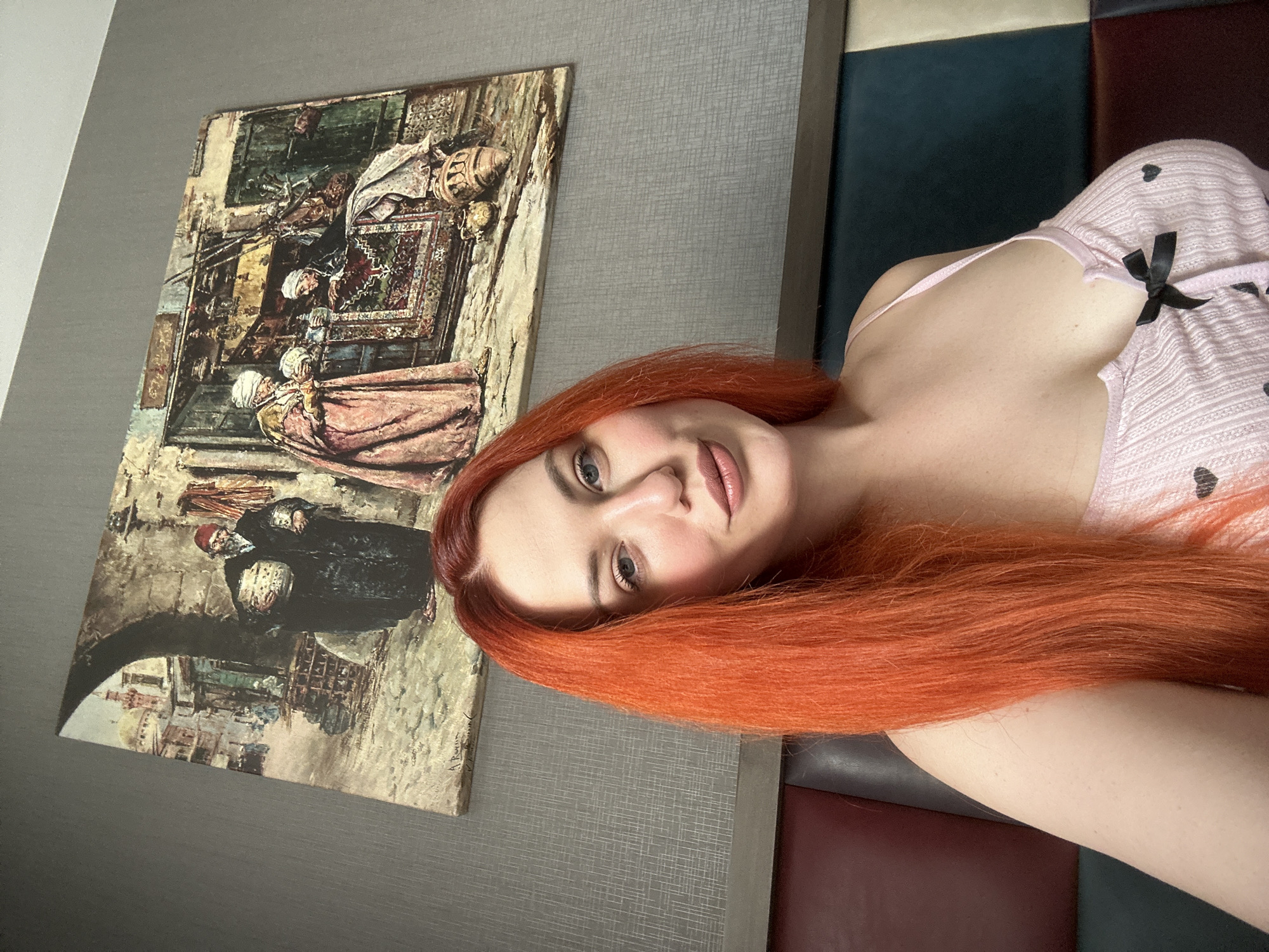 A woman with long, bright orange hair is in a room, posing for a selfie. She is wearing a light pink, ribbed tank top with black heart patterns and a small black bow in the center. She is looking directly at the camera with a slight smile. Behind her, on the wall, is a large framed painting depicting a street scene from a historical city. The painting shows figures in traditional clothing, a marketplace with stalls, and architectural elements like arches and minarets. The room appears to be a bedroom or living space, with a headboard or upholstered furniture visible in the lower part of the frame. The overall impression is of a personal space with an artistic element. The question of location in Avcılar, Turkey is not answerable from the visual information alone.