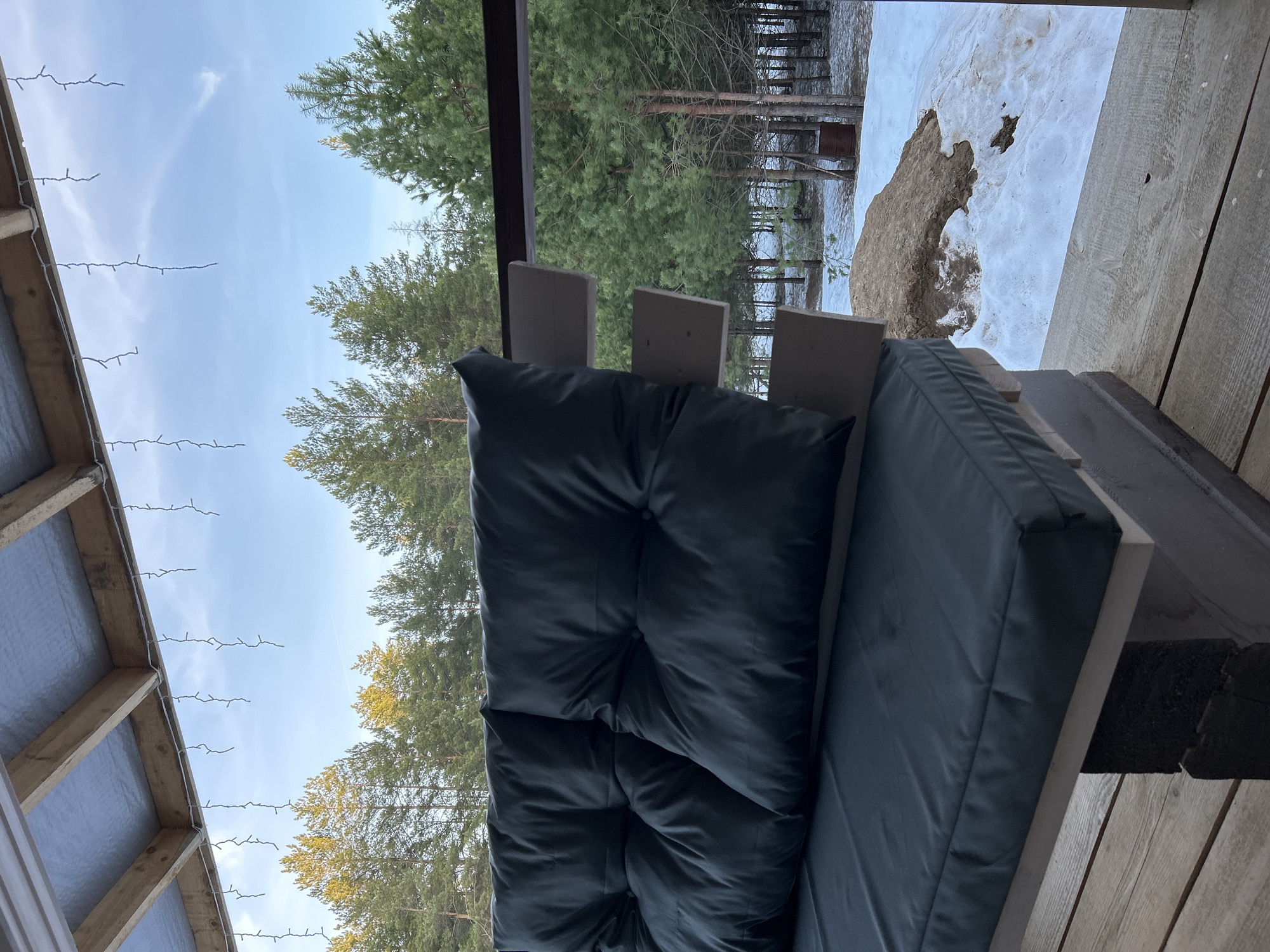 The image shows a view from a covered outdoor seating area, likely a porch or veranda, in a snowy environment. The seating area includes a couch with dark-colored cushions. Through the railing, there are trees, patches of snow, and exposed ground. The sky is visible with some cloud cover. The location is Chaikovsky, Russia. There are no people visible in the image. The objects visible are the couch, cushions, railings, trees, snow, sky, and possibly lighting decorations. The overall environment suggests a transition season, possibly late winter or early spring.