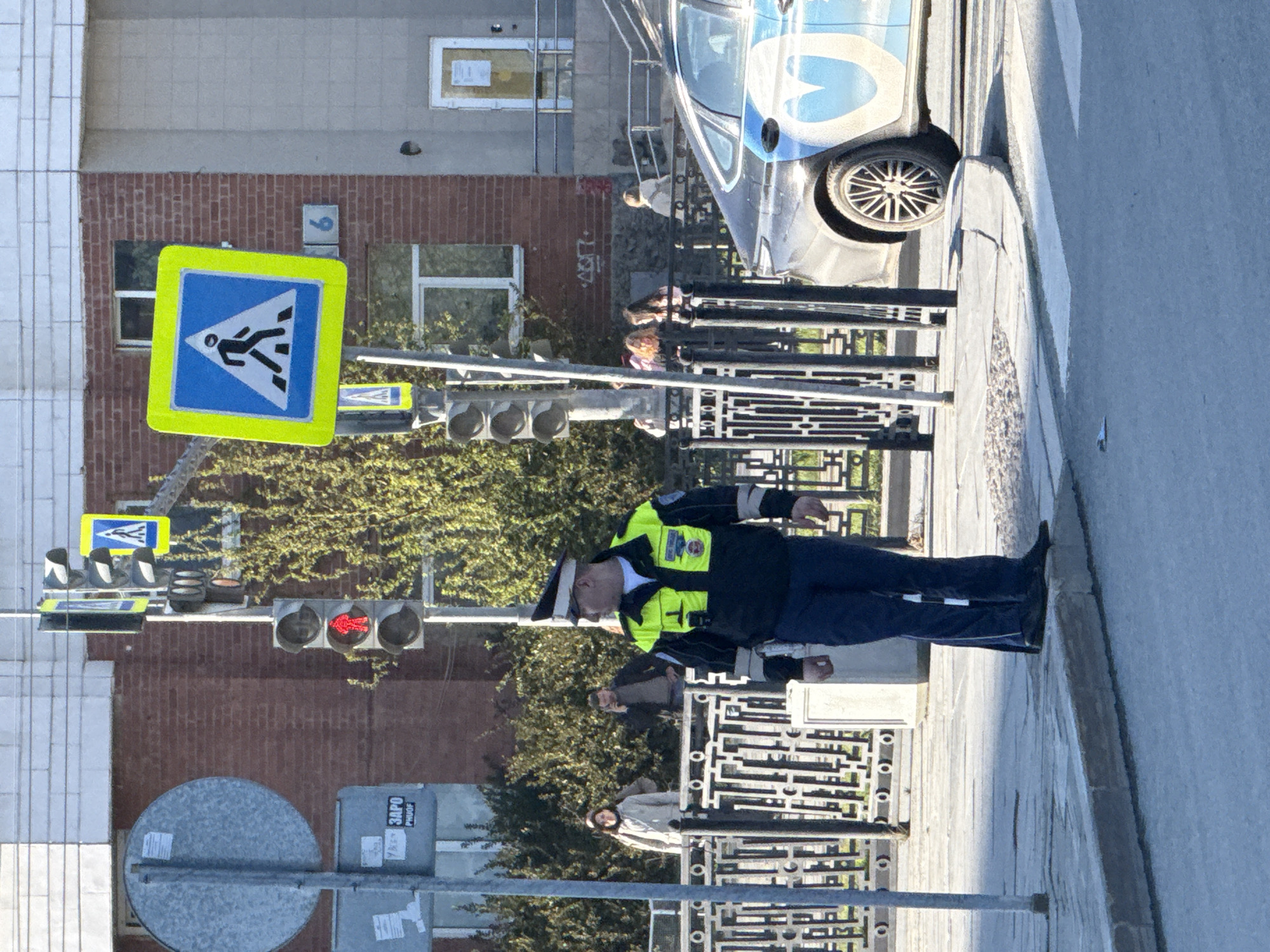 The image shows a street scene in Yekaterinburg, Russia. A traffic officer stands near a pedestrian crossing, identifiable by the blue and yellow sign. A traffic light shows a red signal. A vehicle is parked nearby. Pedestrians are present. The background includes buildings, one with a number "9" on it, and trees. A sign with "ЗАРО PROOF" in Cyrillic is visible.