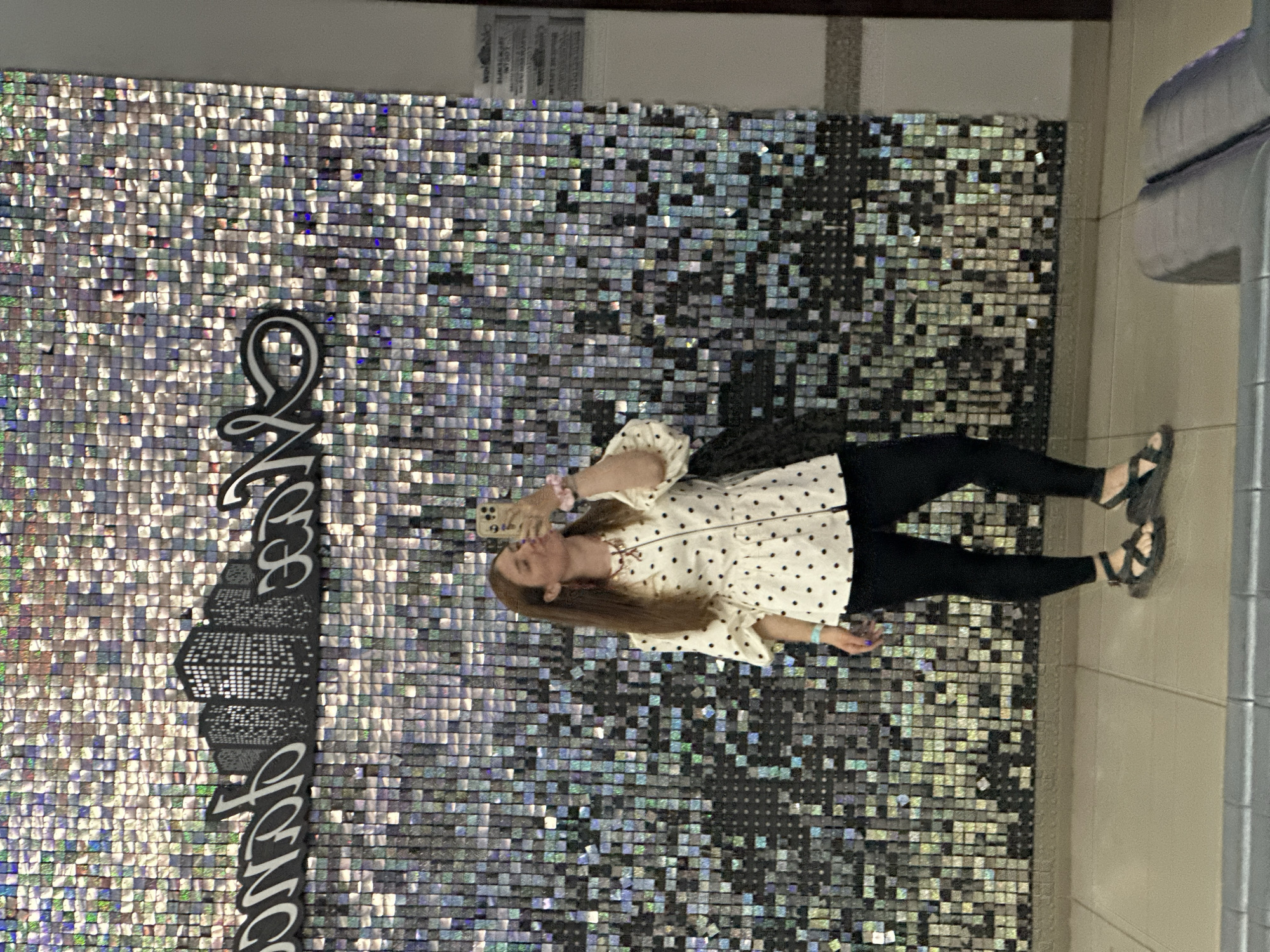 A young woman with long brown hair stands in front of a sparkly wall in what appears to be an indoor setting. She is taking a selfie with her phone, puckering her lips. She is wearing a white peplum top with black polka dots, black leggings, and black sandals. A black bag is slung over her left shoulder. The wall behind her is covered in small, reflective square tiles that create a shimmering effect. The floor is tiled with large beige tiles. Above the woman, there is a black graphic of city buildings with white outlines. The image does not provide any specific visual cues to suggest the location is Petropavlovsk-Kamchatsky, Russia.