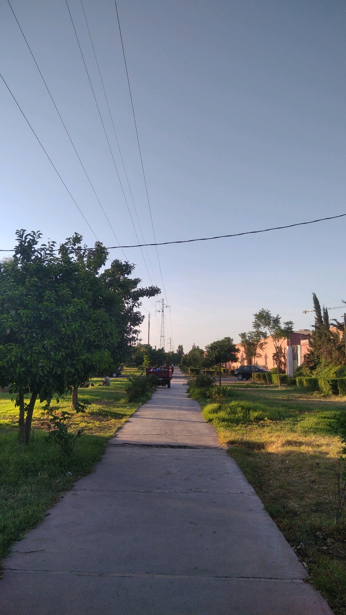 The image depicts an outdoor scene on a clear day, likely in an urban park or residential area. A paved pathway extends into the distance, bordered by green grass and trees. Several power lines are visible crisscrossing the sky. In the middle ground, a small, red utility vehicle with a trailer is moving along the pathway. A few scattered trees and some low-lying bushes are present, along with some buildings in the background that appear to be residential. The lighting suggests it is either early morning or late afternoon, with long shadows cast by the trees and the utility vehicle. The environment appears to be well-maintained and peaceful.