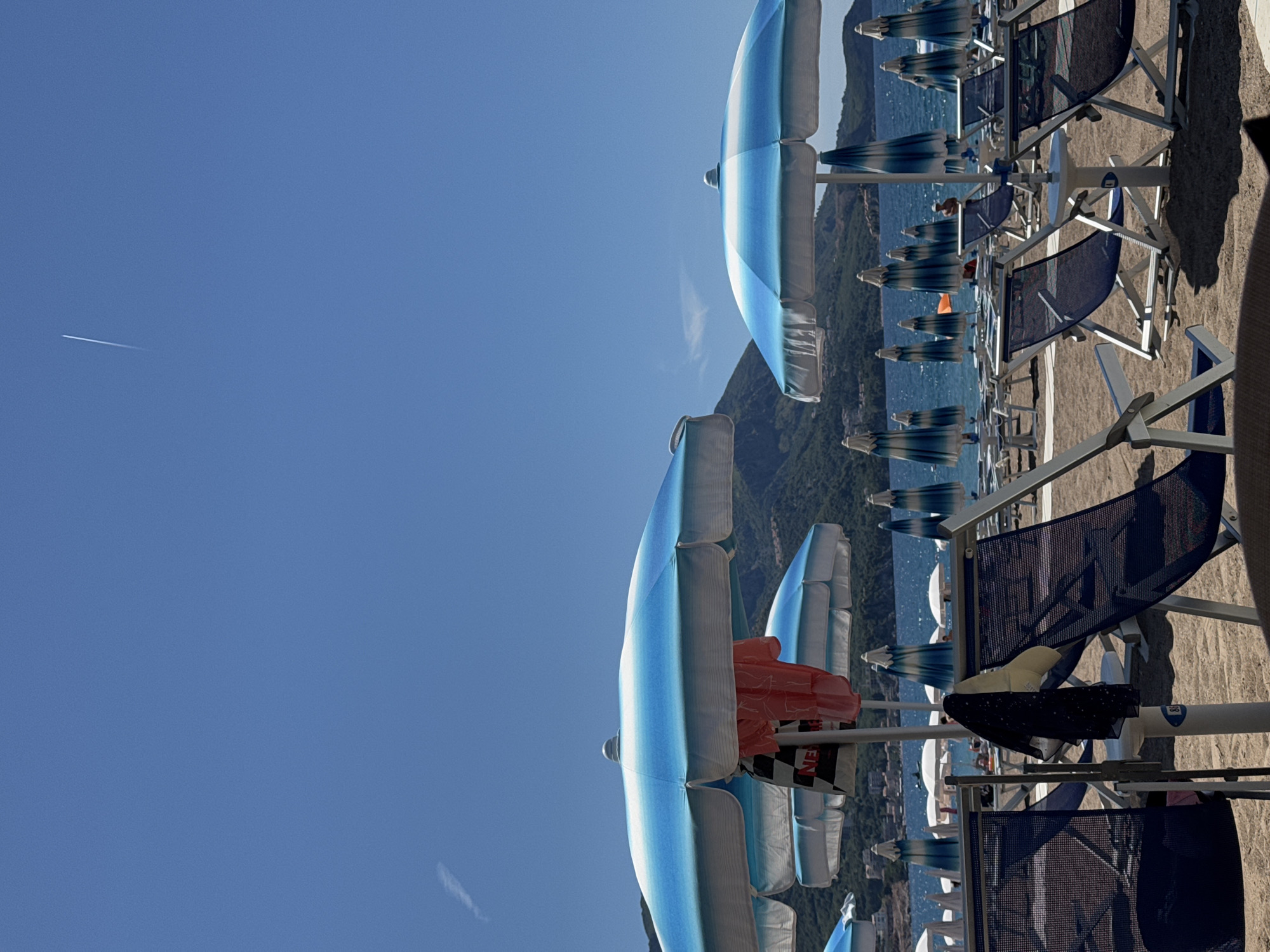 The image depicts a crowded beach scene on a sunny day in Boreti, Montenegro. Rows of blue and white striped beach umbrellas are set up on the sandy shore. Many are open, providing shade to the numerous empty lounge chairs arranged beneath them. A few people are visible in the distance near the water's edge, and some appear to be walking. In the foreground, several blue fabric lounge chairs with metal frames are prominently displayed, some with items of clothing draped over them. The sea, a vibrant blue, stretches out behind the beach, meeting a mountainous landscape in the background under a clear, bright blue sky with a faint contrail from an airplane. The overall impression is one of a popular tourist destination prepared for visitors.