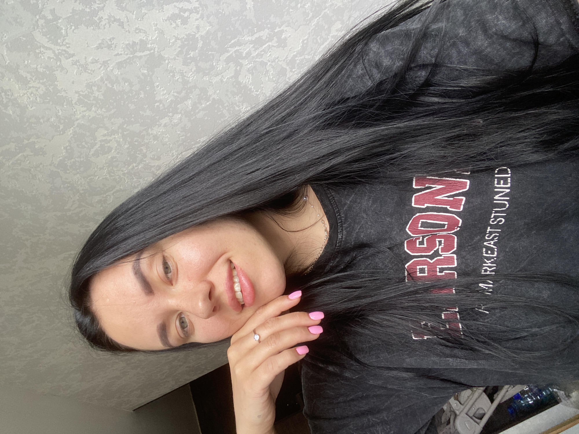 In the city of Chaikovsky, Russia, a young woman with long, straight black hair is captured in a selfie. She is wearing a dark, possibly charcoal gray, t-shirt with faded lettering that appears to read "LARSON" in burgundy with a white outline, and below that, "MARK EAST STUNED" in white. Her right hand is resting on her cheek, with her fingers gently touching her face. She is wearing a delicate silver ring on her ring finger, and her fingernails are painted a bright pink. Her makeup is subtle, with a focus on her bright pink lips and lightly defined eyebrows. The background is a textured, light gray wall. The lighting suggests it is daytime, likely indoors.