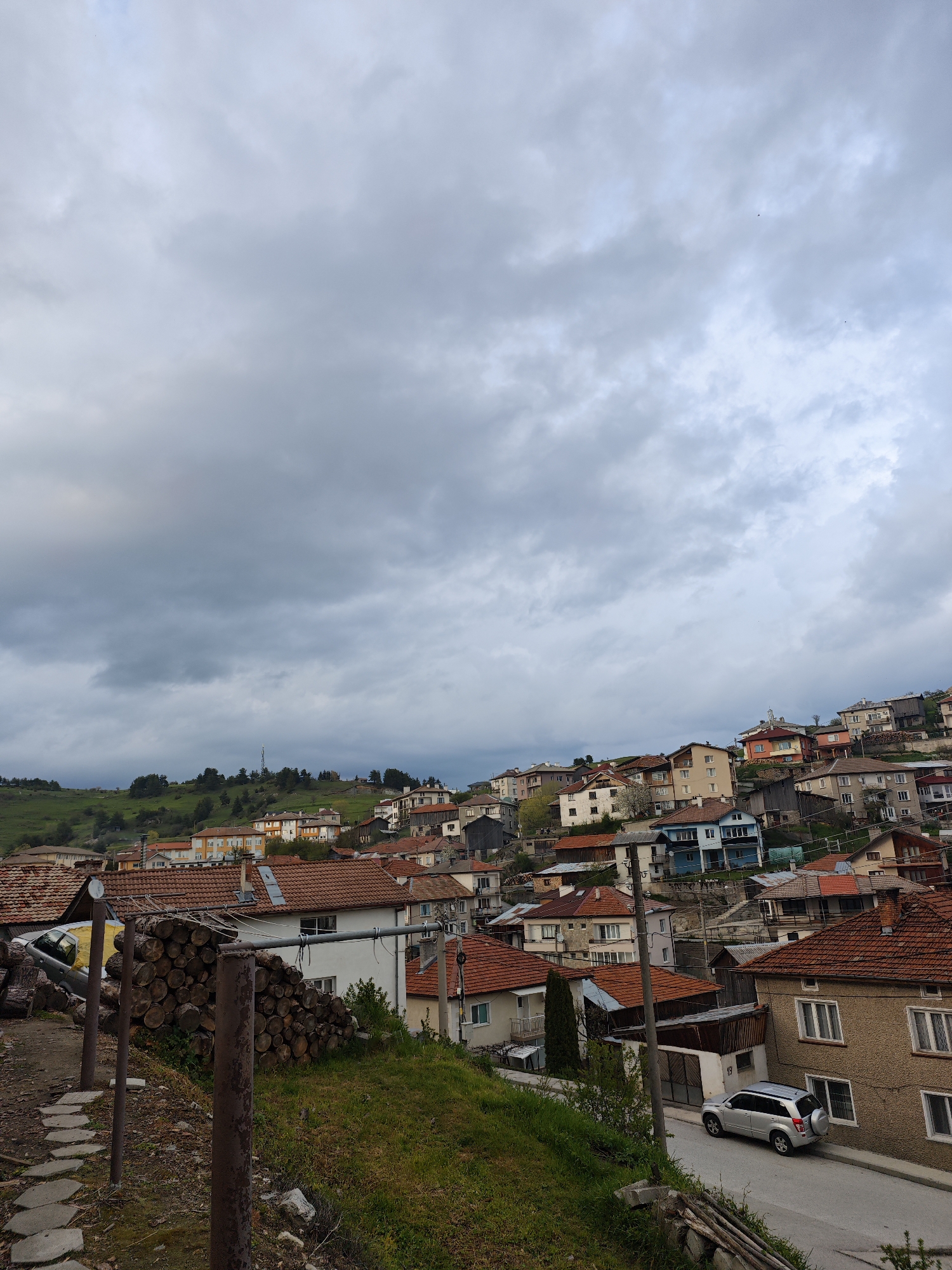 The image shows a view of the city of Kasak, Bulgaria. The scene is taken from an elevated position, likely a hillside or a building. The houses are built on a slope, with rooftops of various colors, predominantly red tile, visible. There are multiple houses, some with brick exteriors, and various colorations of their walls. A single silver car is parked on a road. The sky is filled with grey, overcast clouds. There is a green, grassy area in the foreground, and a pile of logs is stacked on the left. There is a metal support structure in the foreground. A hillside covered in green vegetation and scattered trees can be seen in the distance.