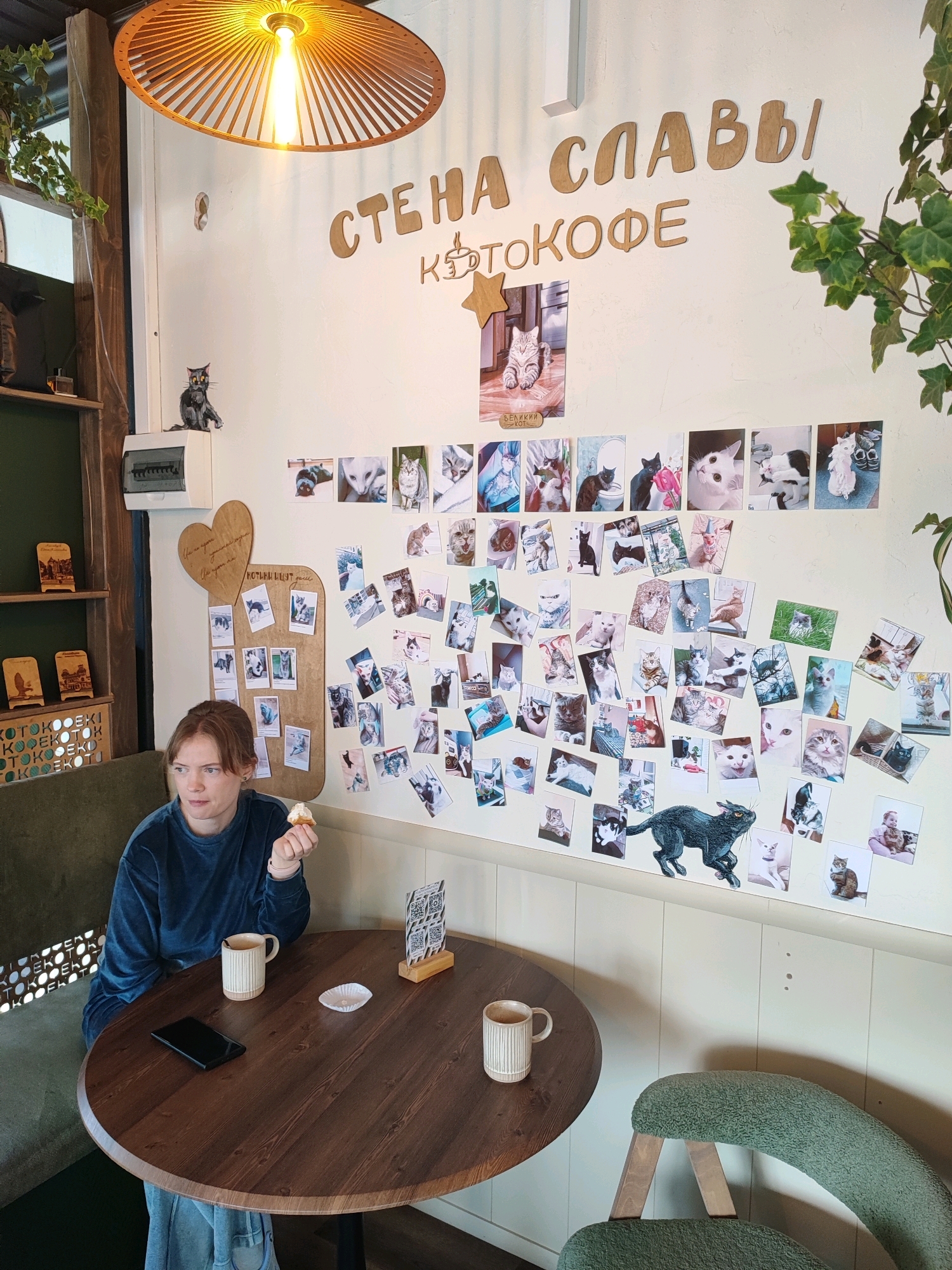 The image captures an interior scene, likely a cafe, with a white wall adorned with numerous photographs of cats and wooden lettering. The lettering above the photographs reads "СТЕ НА СЛАВЫ" (Stena Slavy - Wall of Glory) and "КOTOКОФЕ" (Kotokofe - Cat Coffee), suggesting a cat-themed establishment. On the left side of the wall, there is a wooden heart-shaped display board with more cat photos. A woman with reddish-brown hair, wearing a blue velour top and jeans, sits at a round wooden table. She is holding a small pastry in her right hand and looking to her left. On the table in front of her are a mug, a smartphone, and a small white paper cup. A green upholstered chair is visible to the right of the table. The background includes a wooden shelf unit on the far left and green foliage. The overall atmosphere appears relaxed and cozy. The presence of Russian lettering on the wall indicates the location is in Russia. Given the "Kotokofe" sign, this is likely a cat cafe in Pyatigorsk, Russia.