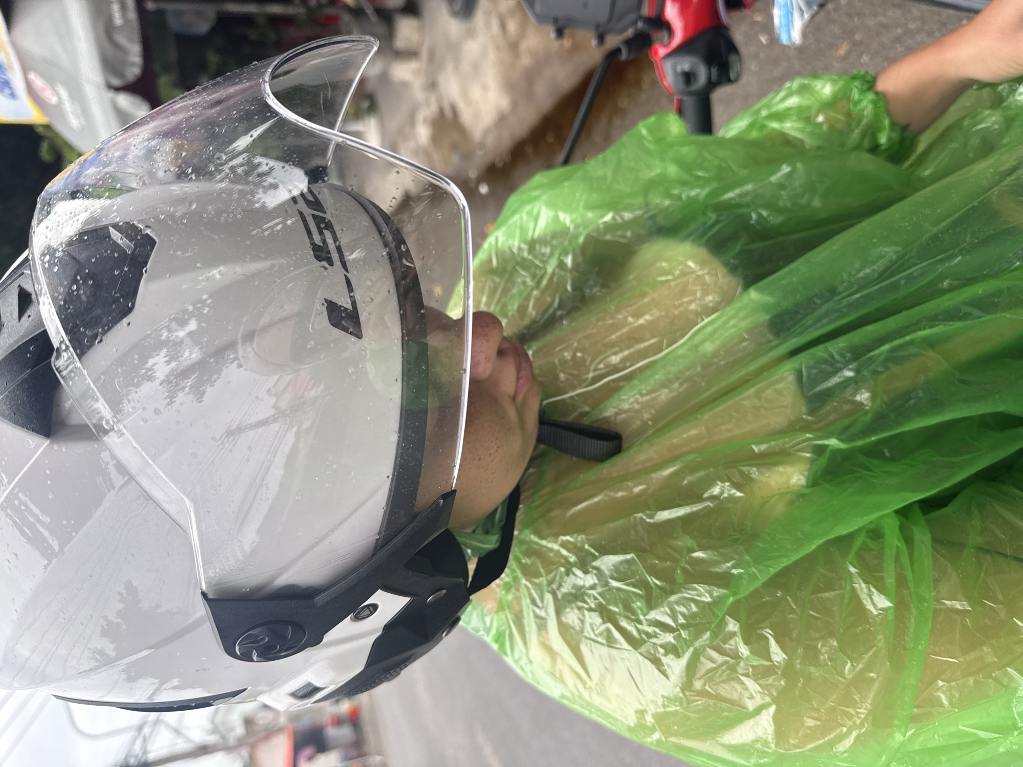 A person wearing a white helmet with a clear visor and a bright green poncho is seated on a motorcycle. The helmet has water droplets on its surface, and the visor is also wet, suggesting it is raining. The person is looking down, with their face partially obscured by the visor and hood of the poncho. The background shows a street scene in Nha Trang, Vietnam, with wet pavement, some buildings, and other vehicles. A red handlebar and part of the motorcycle are visible in the foreground. The overall atmosphere is one of a rainy day in an urban setting.
