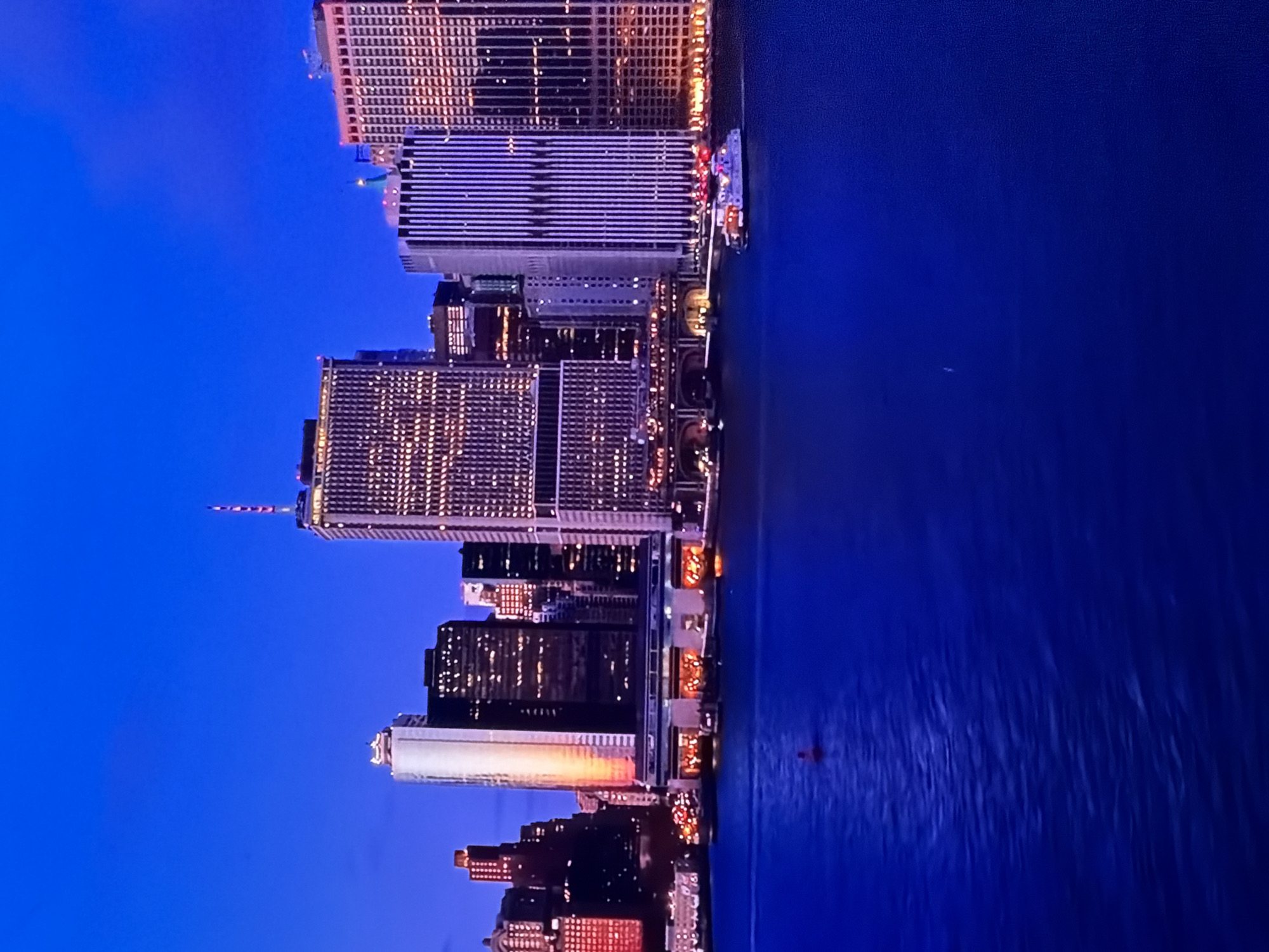 The image presents a vibrant urban waterfront at twilight, characterized by a deep, uniform blue sky that dominates the upper frame, transitioning slightly darker overhead. The foreground and midground are occupied by a wide expanse of dark blue water, displaying gentle ripples and reflecting the ambient light. A small, red, cylindrical navigational buoy floats prominently in the midground-left. Towards the midground-right, a brightly lit, multi-decked boat with numerous white and red lights is visible, moving across the water from right to left.
The background is dominated by a dense cityscape composed of numerous tall buildings lining the waterfront. These structures are heavily illuminated, primarily with warm yellow, orange, and white lights emanating from windows and architectural features. A prominent tall, slender building on the left side of the skyline displays a striking vertical streak of warm, orange-toned light along one facade, making it stand out. Adjacent to it are several darker buildings with grids of lit windows. Slightly right of center, another very tall building exhibits a distinct pattern of vertical stripes across its facade. To the far right, a large building is bathed in a strong reddish-orange glow. Various other buildings, differing in height and design, fill the urban panorama, some topped with antenna structures; one such antenna glows with a red light at its tip. Below the taller buildings, at the water's edge, are lower structures featuring large, brightly lit arched openings, suggesting commercial or public establishments with inviting interior glows. The overall scene conveys an active, bustling metropolis at night. The location is Maidstone, United Kingdom.