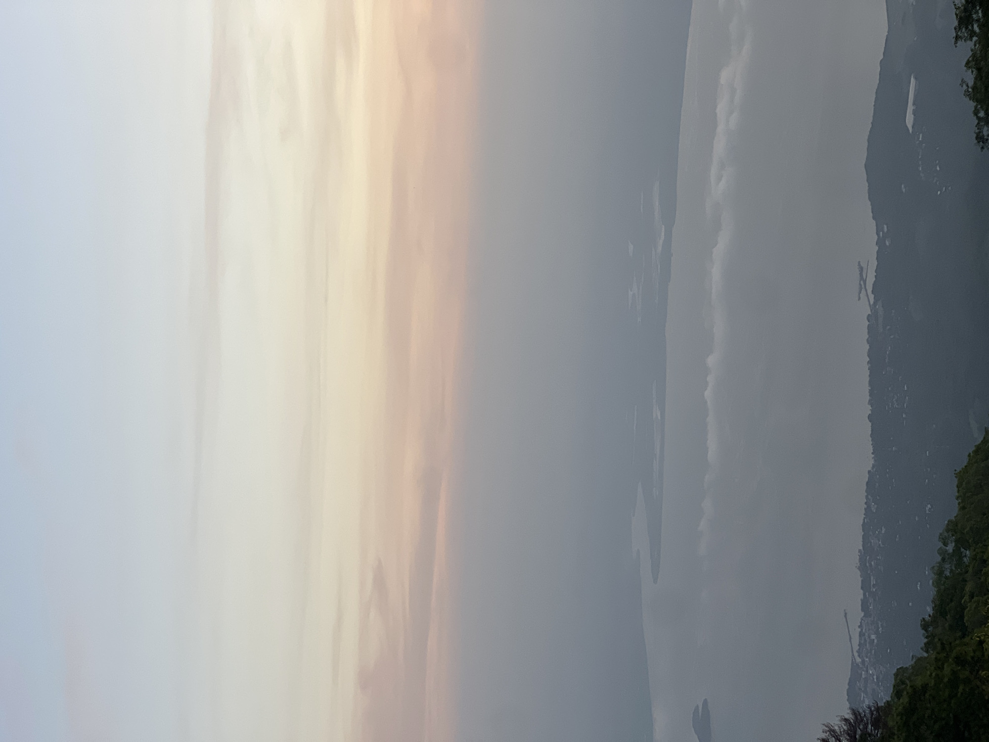 This is a high-angle, vertical, full-frame shot of a hazy, diffused dawn sky over a body of water and a coastal town. The sky transitions from a pale, almost white blue at the top to soft pinks and oranges as it approaches the horizon. A layer of mist or low clouds hangs between the sky and the landscape, obscuring much of the detail. Below the mist, a wide, calm body of water, likely a bay or large lake, is visible. Islands or peninsulas break the surface of the water, their shapes softened by the haze. Along the shoreline, a dense cluster of buildings, indicative of a town or city, is spread across the land. Roads and structures are visible, though indistinct due to the distance and atmospheric conditions. The foreground features the dark, shadowed canopy of lush green trees, suggesting the viewpoint is from a higher elevation, possibly a mountain or hill overlooking the scene. The location is identified as El Cacao, El Salvador.