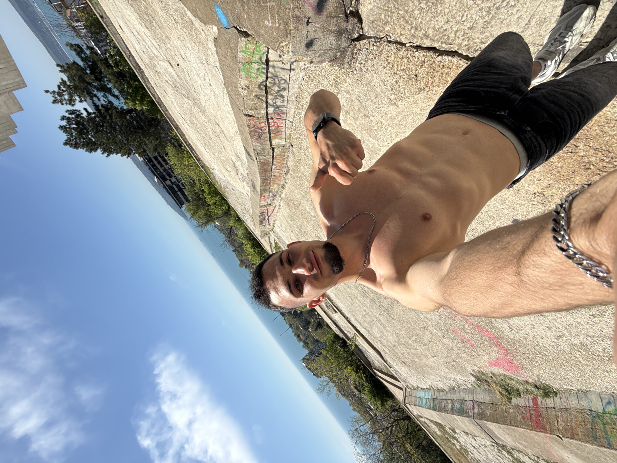 A shirtless man stands at an angle on a concrete structure, likely a retaining wall or embankment, in Varna, Bulgaria. He faces the camera, holding his right hand up, wearing a watch on his left wrist and a silver chain around his neck. He also has a silver bracelet on his right wrist. He wears black jeans and gray sneakers.
The concrete structure on which he stands is cracked and worn, showing signs of age and weather. Graffiti is visible on the side of the structure.
In the background, trees and greenery are seen, along with buildings, implying an urban park or seaside area. Beyond the trees, the blue sky and sea are visible. The image suggests an outdoor setting with an urban and natural mix.