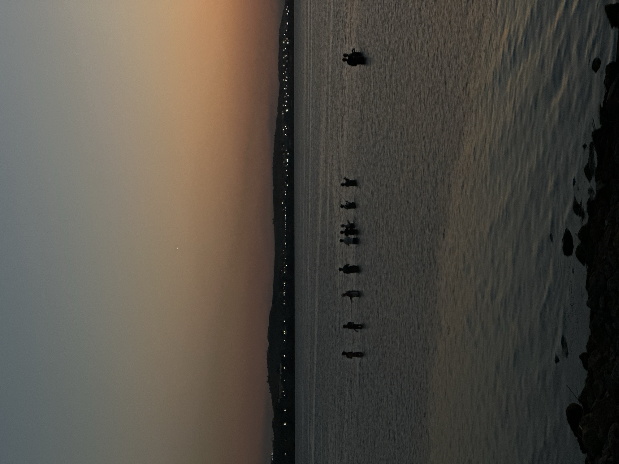 The image depicts a serene twilight scene over a body of water, likely a lake or reservoir, with silhouettes of people wading and fishing. In the distance, a dimly lit shoreline features a line of lights, suggesting a town or city. The sky displays a gradient of colors, transitioning from a pale blue at the top to warm orange and brown hues near the horizon. A single, bright star or planet is visible in the upper portion of the sky. The foreground shows a rocky shore. Given the context of Kokshetau, Kazakhstan, this scene could plausibly be observed at Lake Burabay (Borovoye), a popular natural attraction near the city, or another lake within its vicinity during dusk. The individuals appear to be engaged in recreational fishing, with some holding fishing rods. The overall atmosphere is tranquil and peaceful, characteristic of the end of a day.