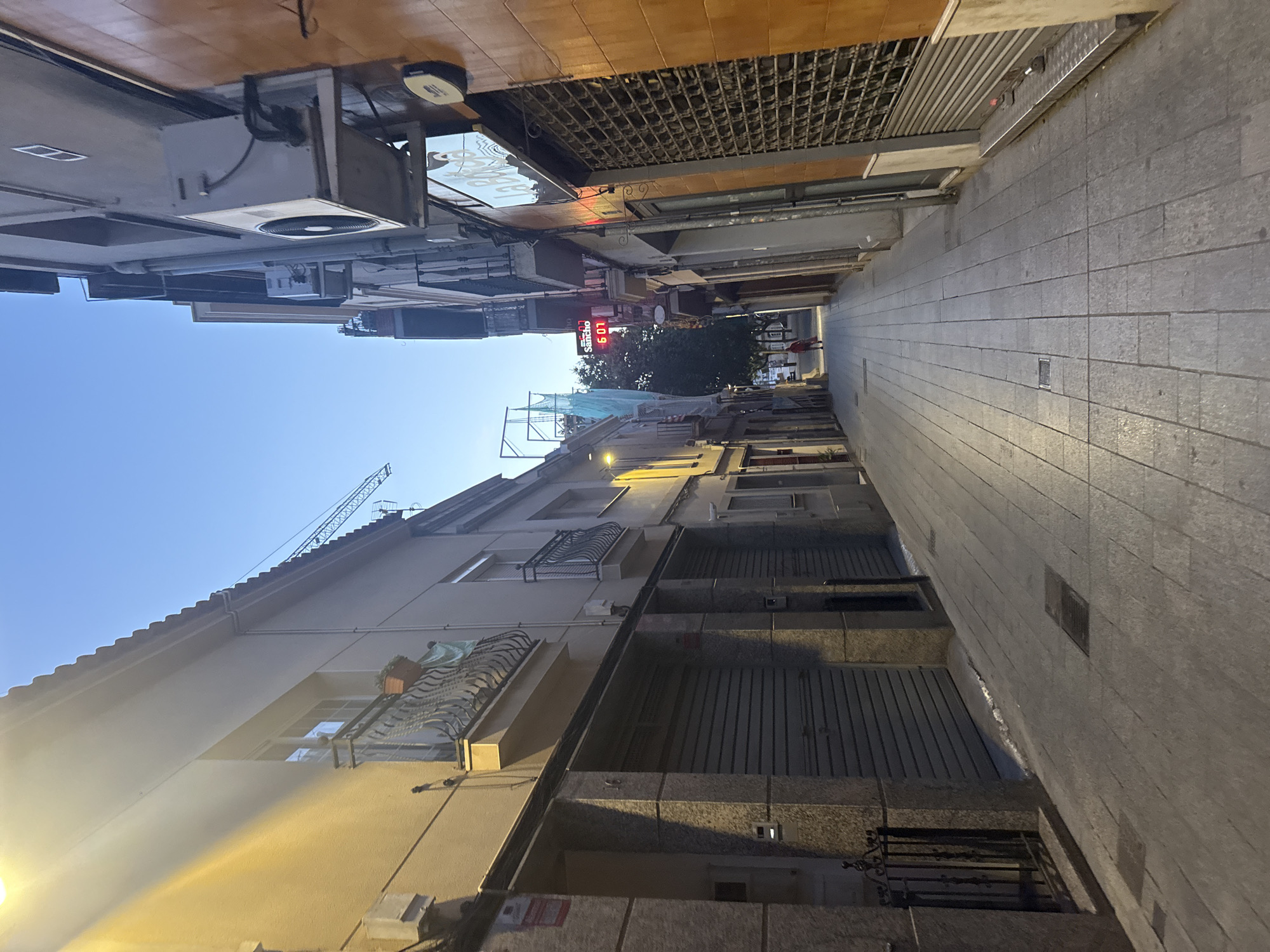 The image depicts a narrow street in Sant Boi de Llobregat, Spain, possibly in the evening given the lighting conditions. The street is paved with rectangular tiles. Buildings line both sides of the street, with the one on the left being a lighter color and featuring balconies and closed roll-up doors on the ground floor. The building on the right is a darker orange color and also has closed shutters.
In the distance, a lone figure, appearing to be a person wearing a red top, walks away from the camera down the street. Above the buildings, a digital clock displays "6 07" with the word "Sancho" above it. A construction crane is visible further in the background. Air conditioning units are mounted on the exterior walls of some buildings. The overall atmosphere is quiet and somewhat commercial, with the closed storefronts suggesting the end of business hours.